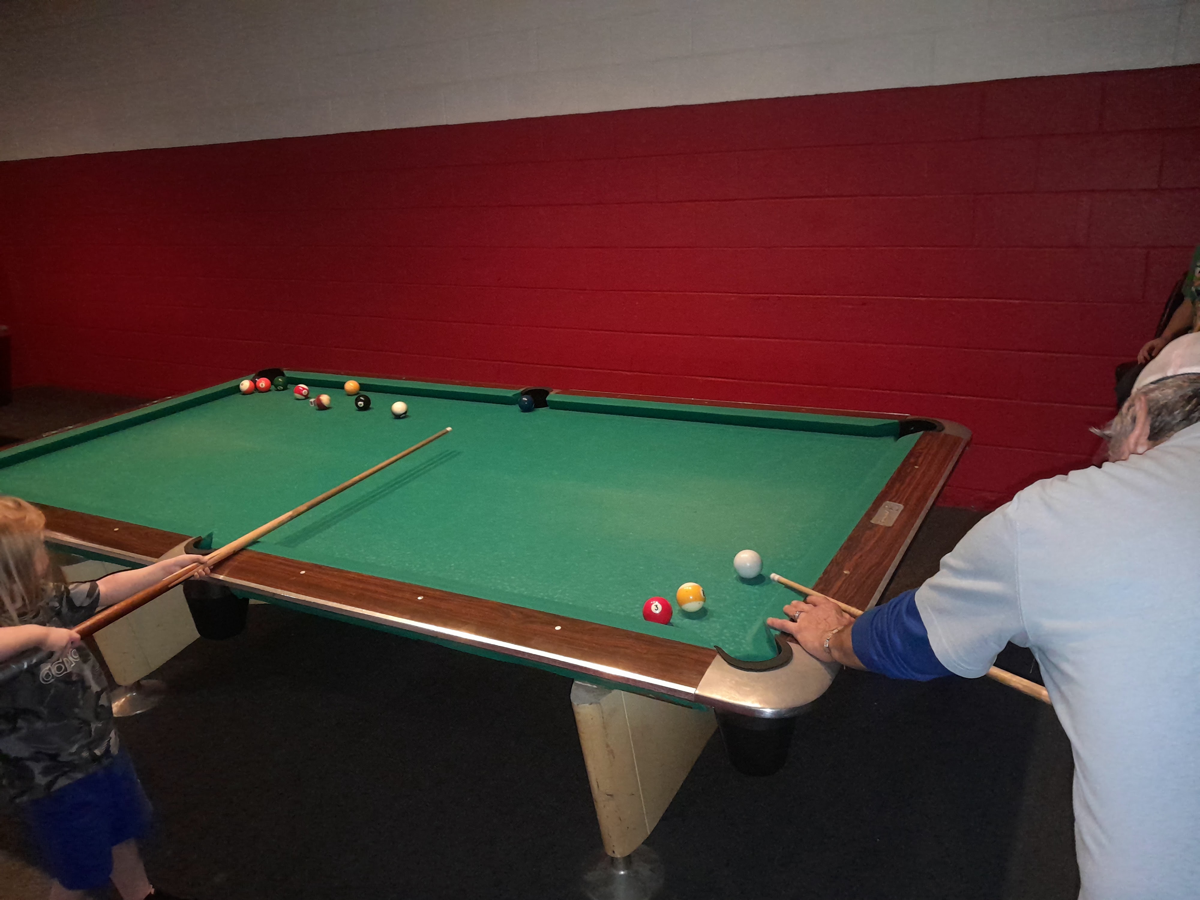 An adult and a small child playing pool on a green billiard table against a red and white wall.