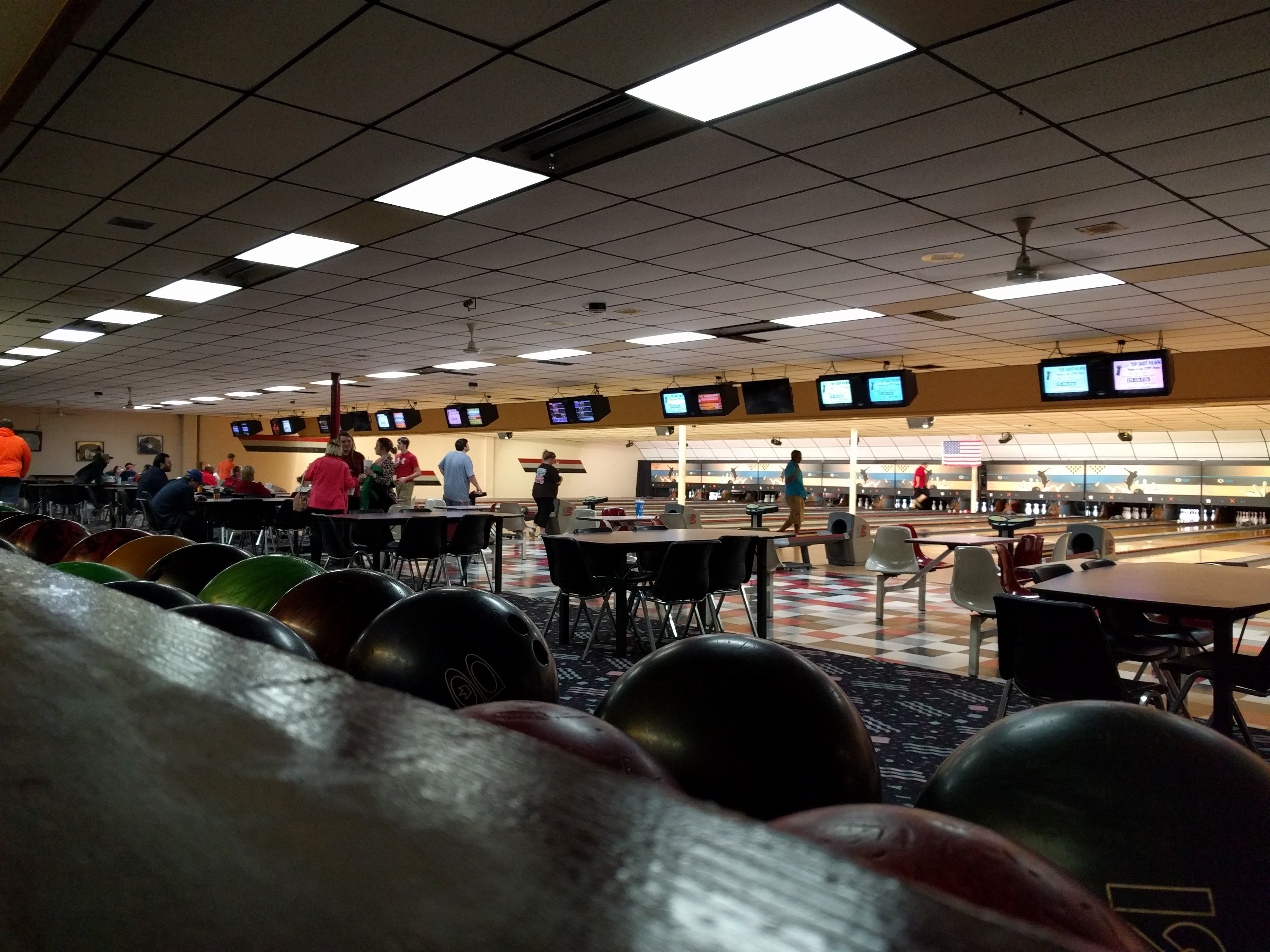 A row of bowling balls on a rack in a bowling center with people and lanes in the background.