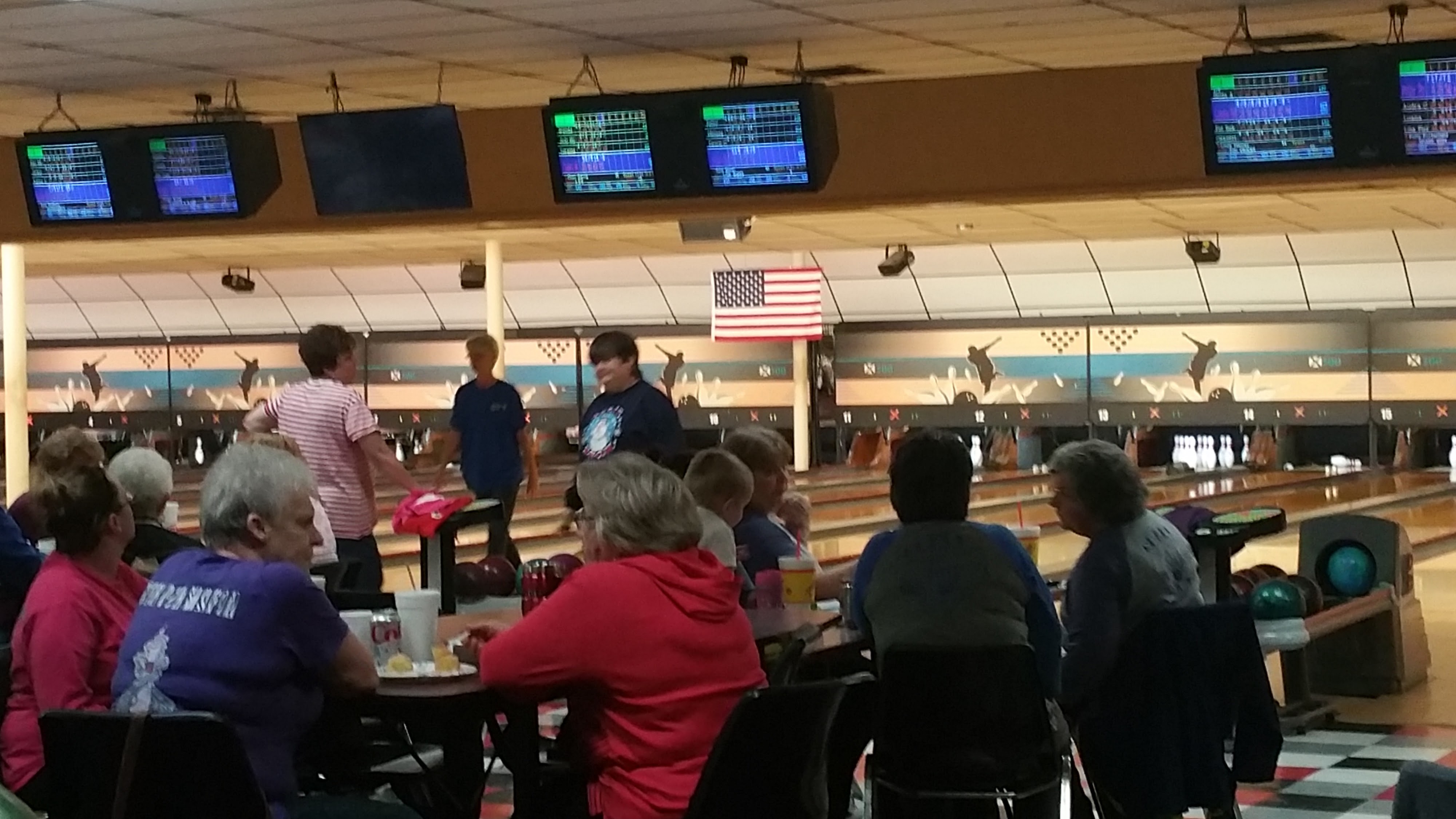 A group of people sitting at tables inside a bowling alley with active lanes and scoring monitors in the background.