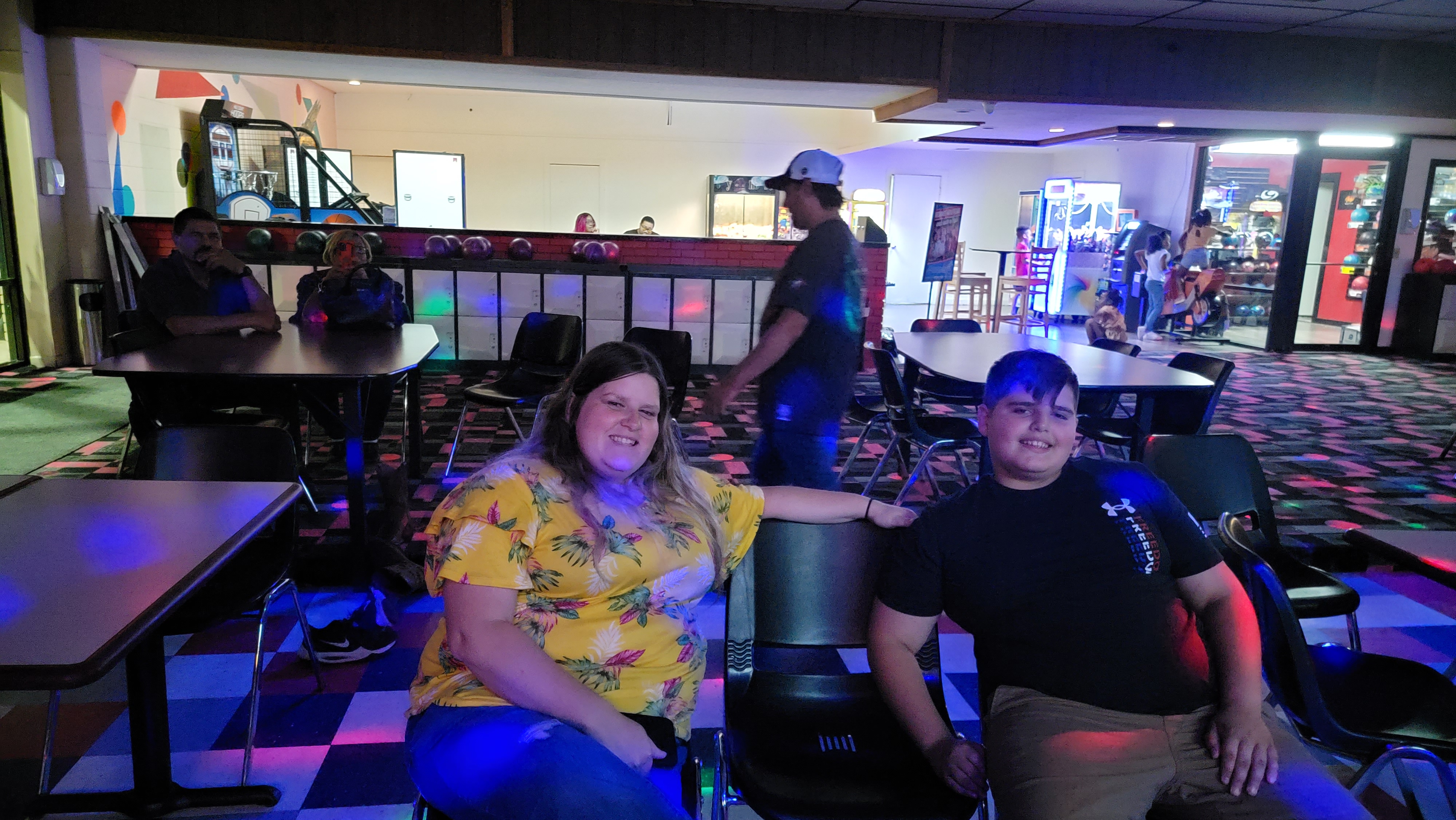 A woman and a young man smiling while seated at a table in a bowling center with colorful cosmic lighting.