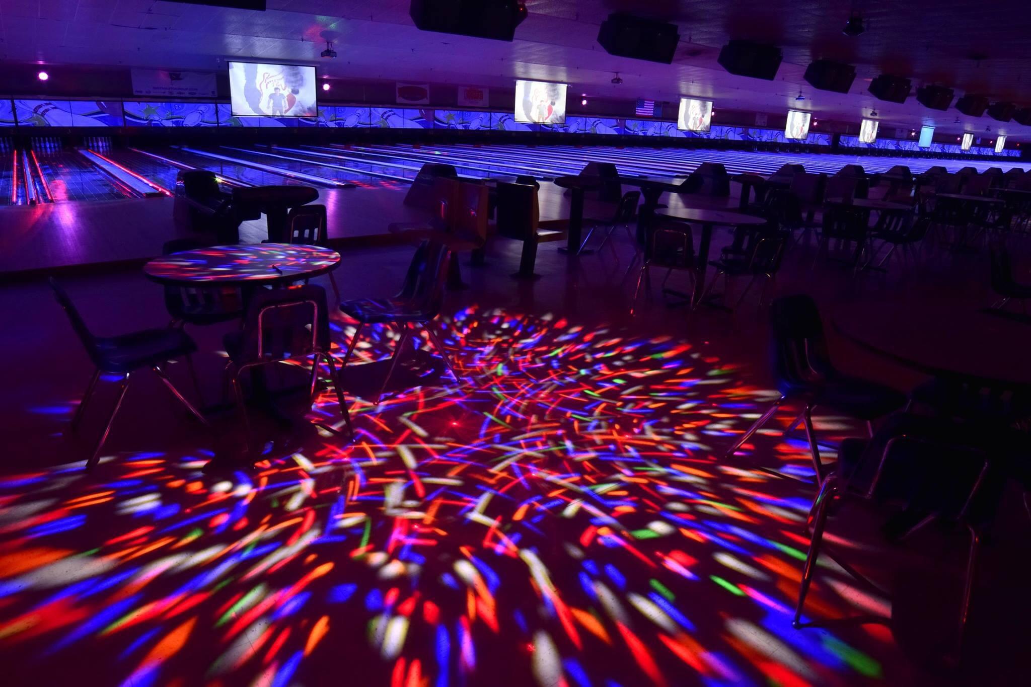 A bowling alley under cosmic lighting, with multiple lanes glowing purple and blue and colorful lights projected on the floor.
