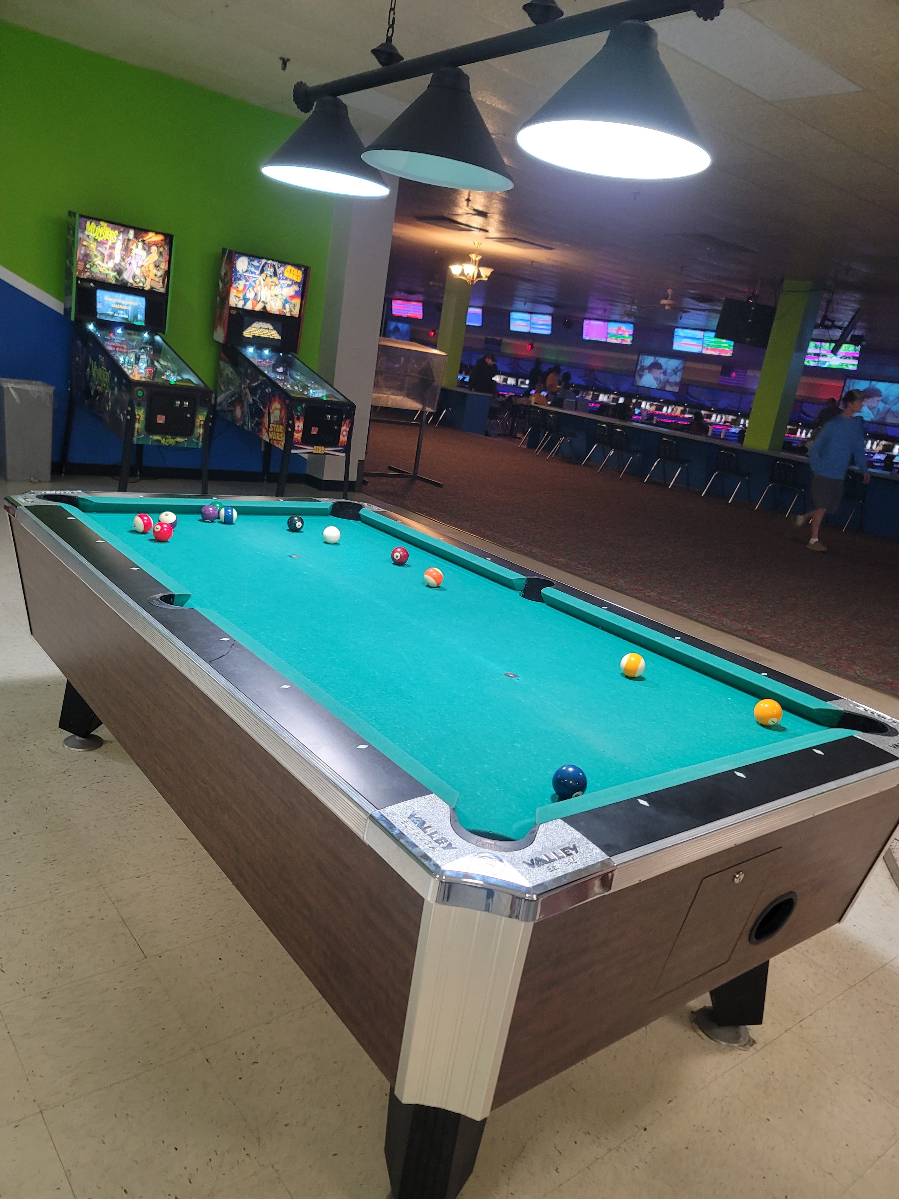 A billiards table with scattered balls in the foreground, with two pinball machines and bowling lanes visible in the background.