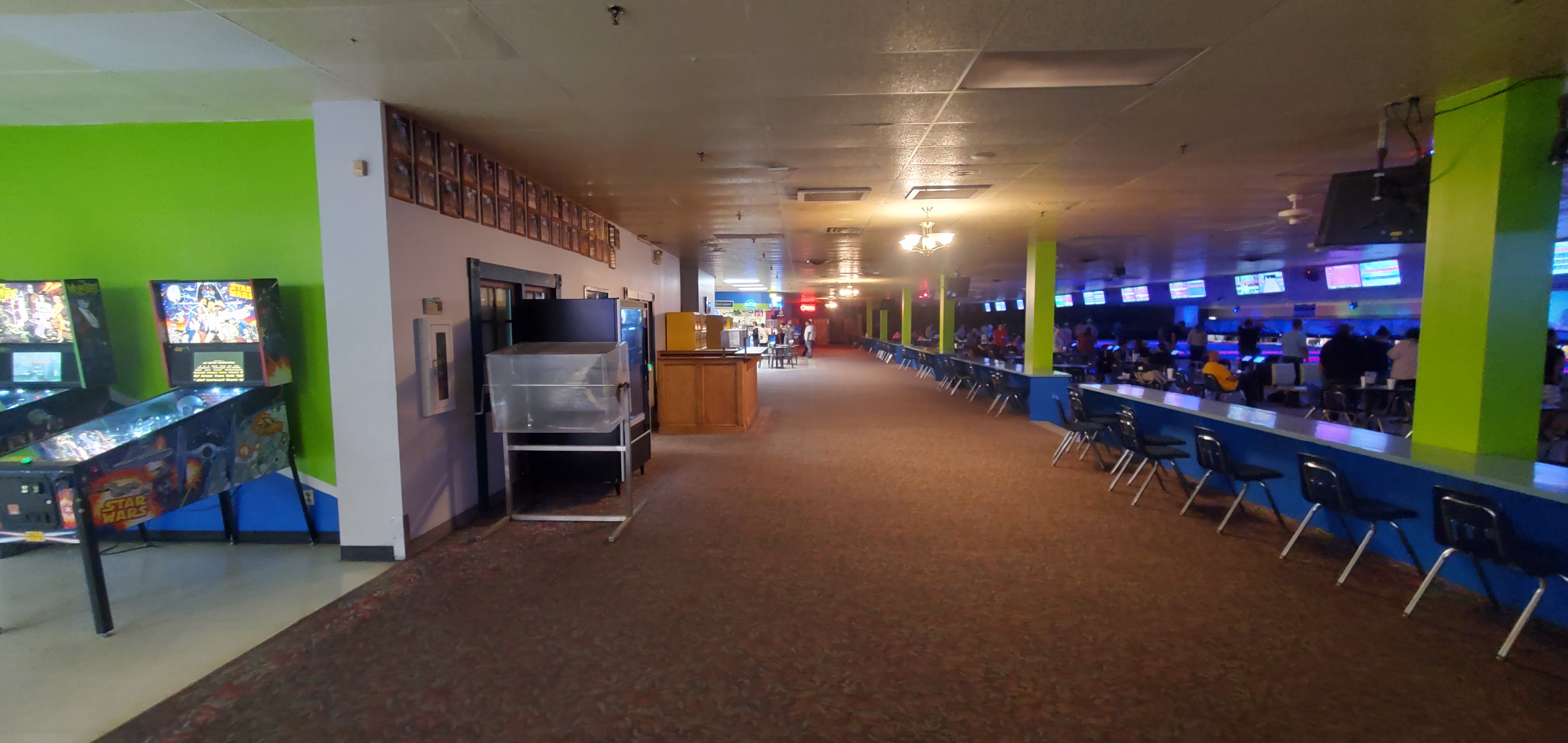A long view down a carpeted hallway leading to active bowling lanes, with arcade games on the left and people bowling on the right.