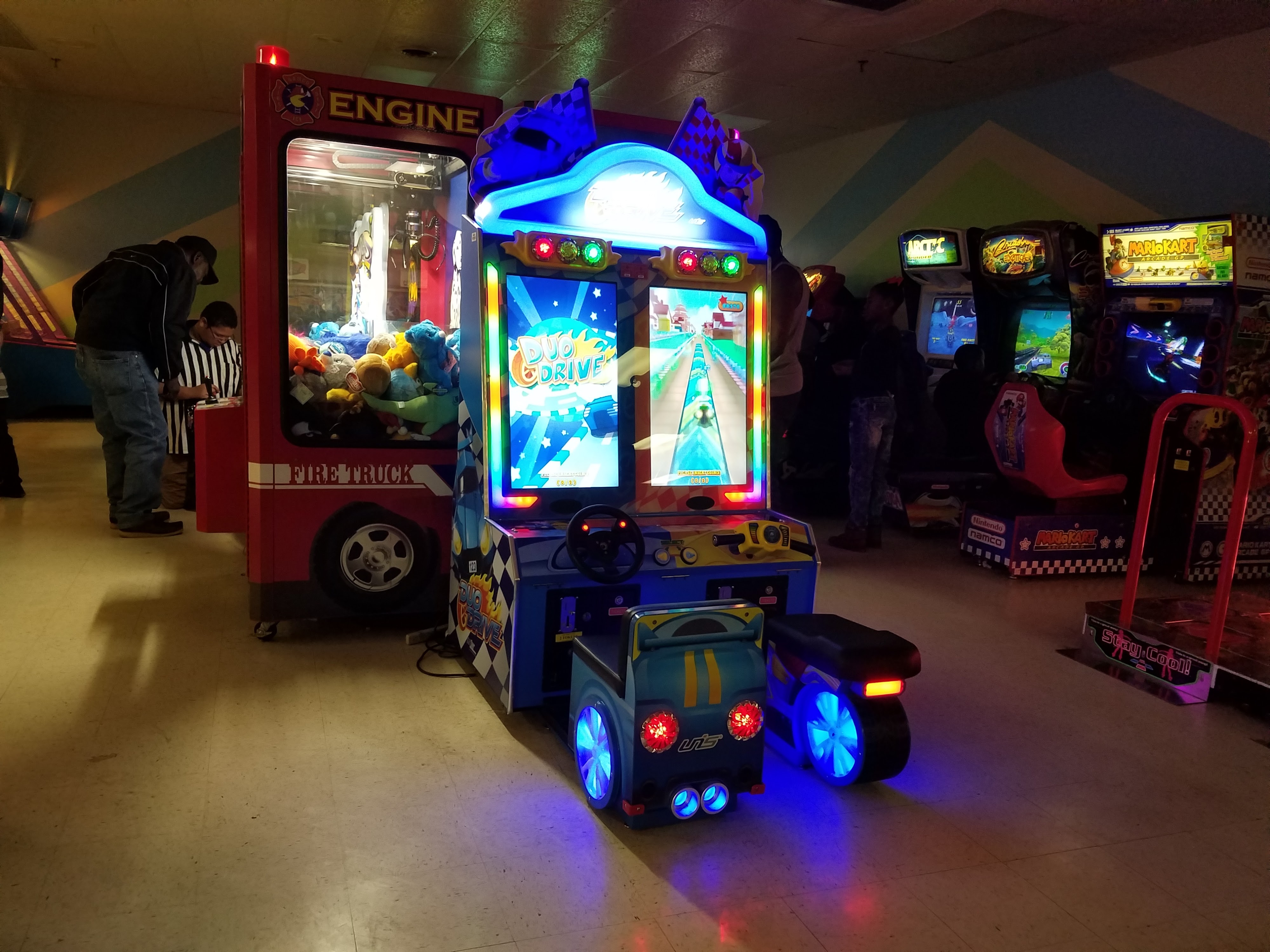 Several arcade games are active in a dim room, including a racing game with glowing wheels and a claw machine.