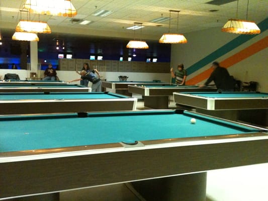 An active billiards room with several people playing pool under overhead lighting, with bowling lanes visible in the background.