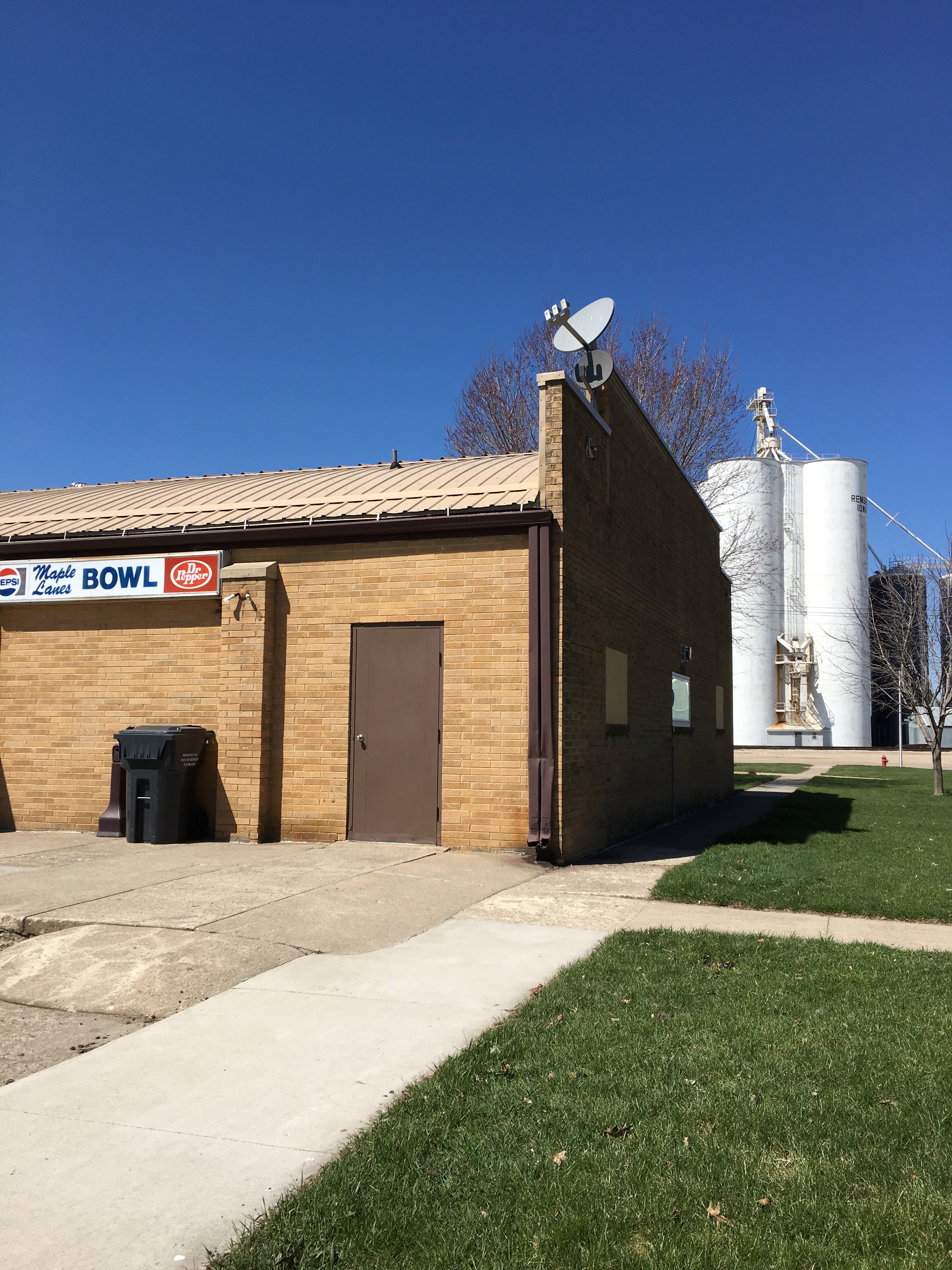 Exterior view of Maple Lanes Bowl building with a clear blue sky, green grass, and grain silos in the background.