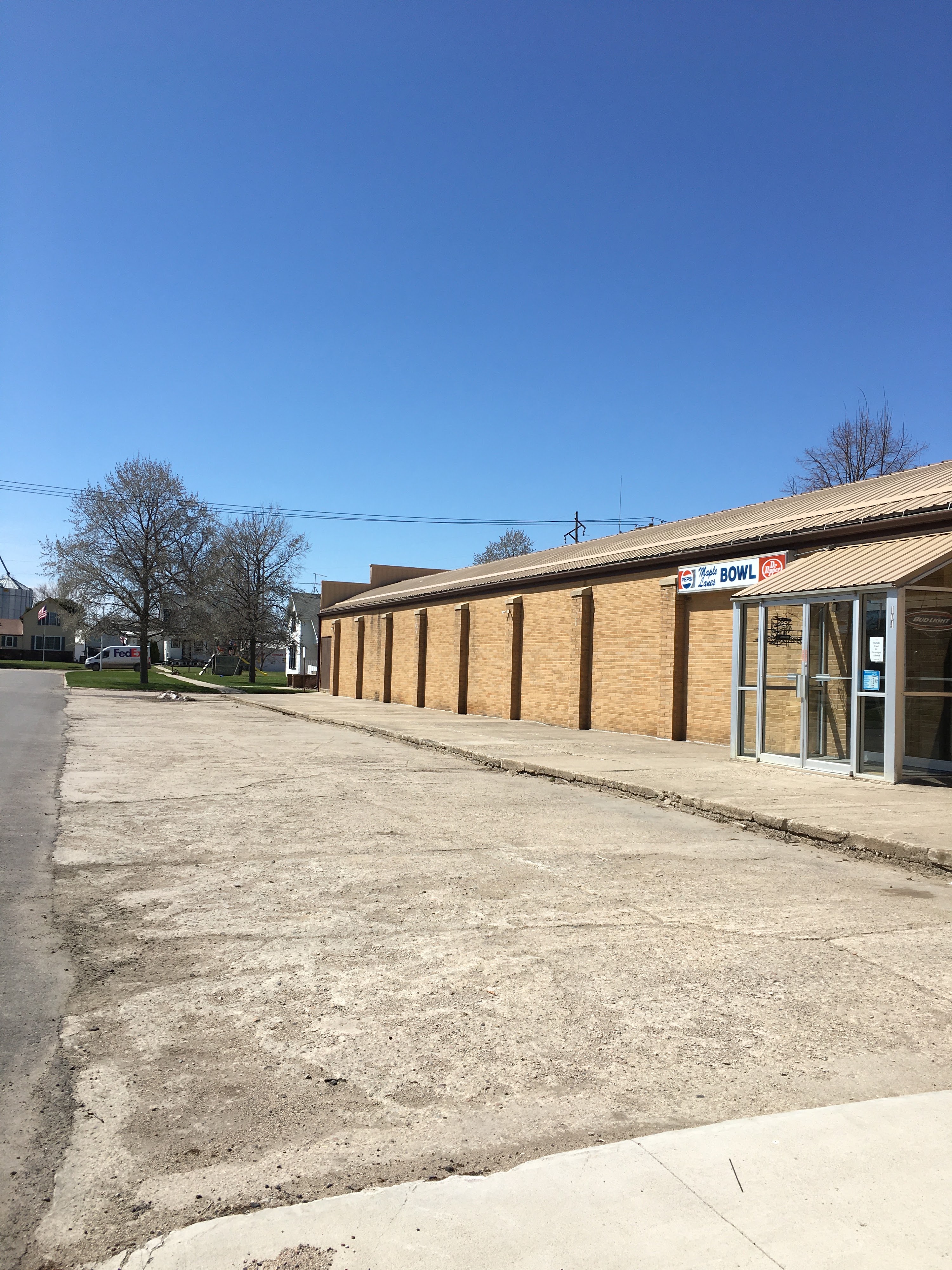 Exterior view of Maple Lanes Bowl building with a tan brick facade, metal roof, glass entrance doors, and an empty parking lot under a clear blue sky.
