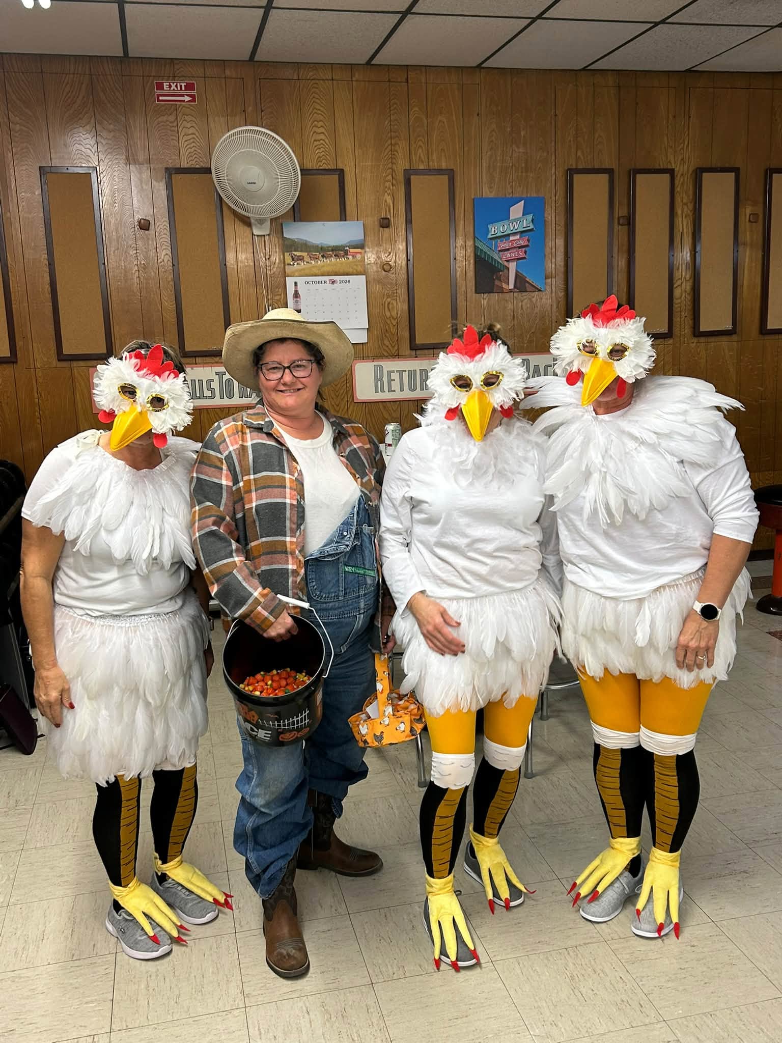 Four people in a bowling center, three in chicken costumes and one in a cowboy hat and overalls holding candy.