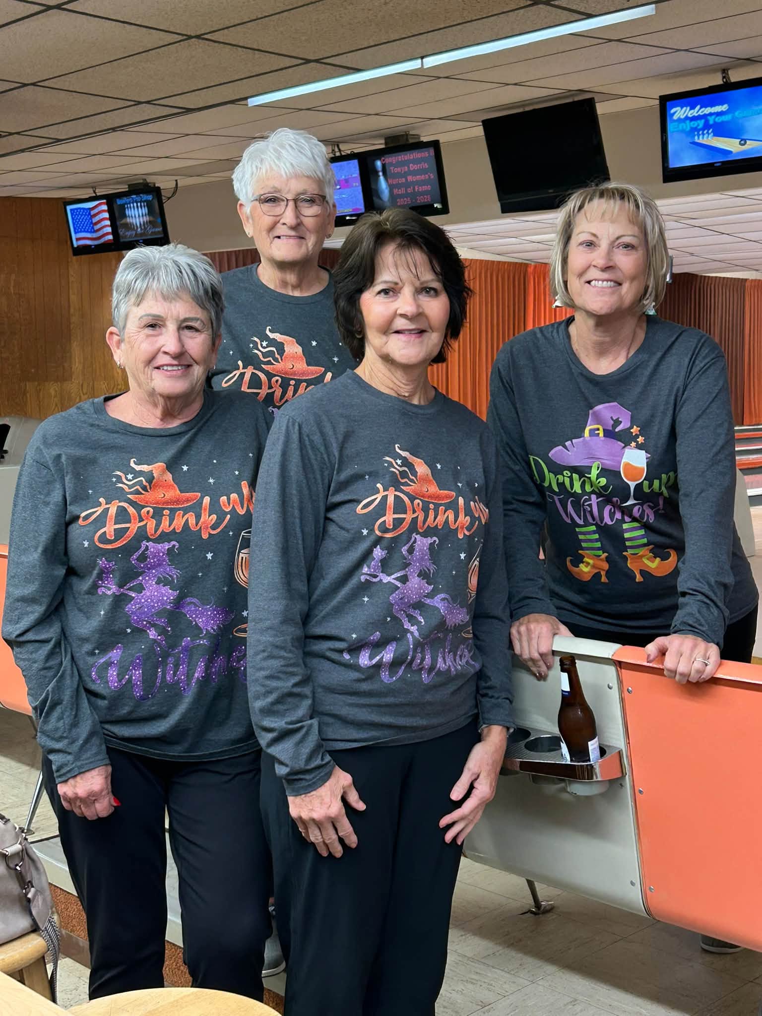 Four smiling women in matching Halloween-themed shirts pose in a bowling alley with lanes and score monitors in the background.