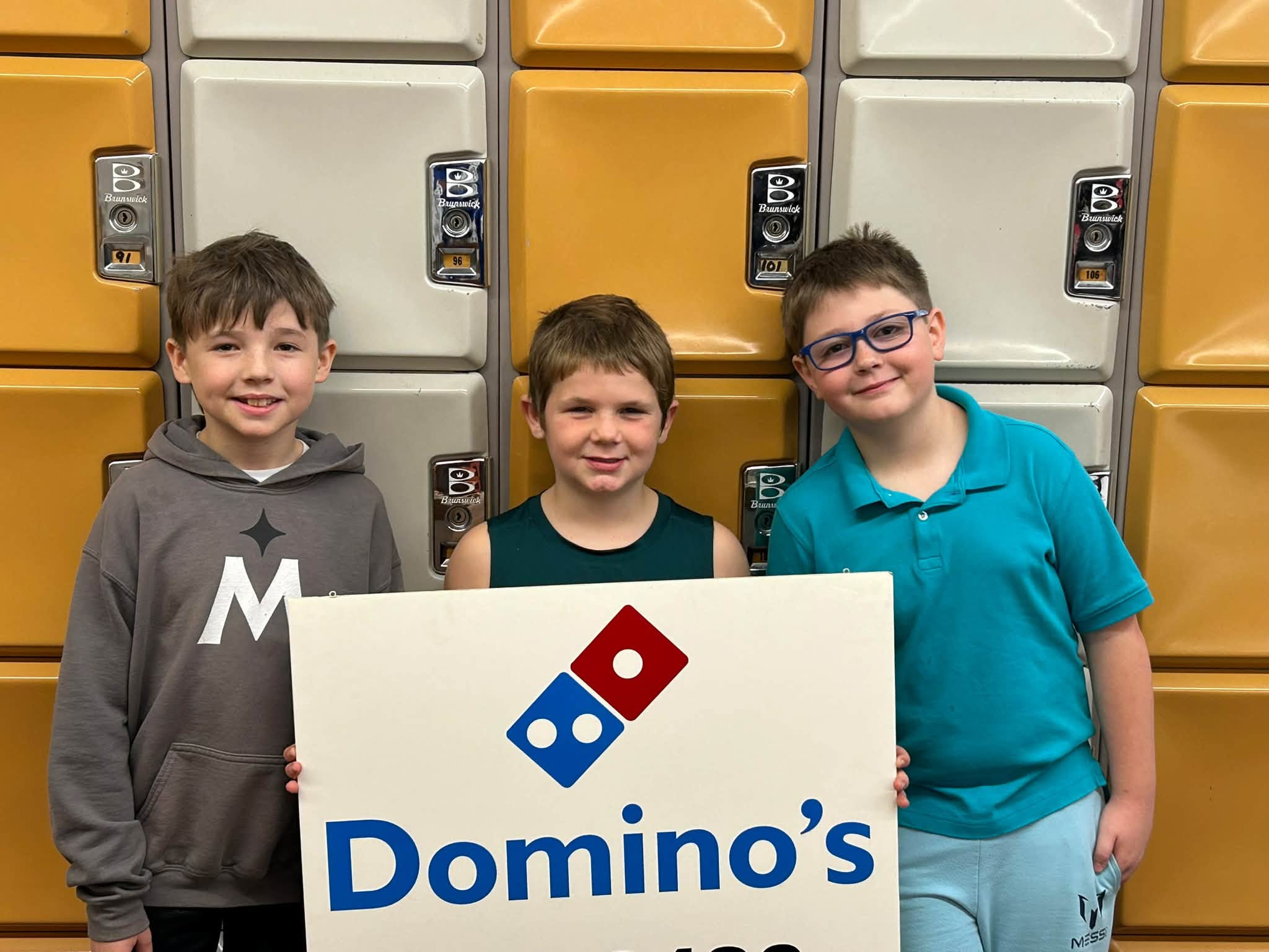 Three young boys smiling while holding a large Domino's Pizza sign in front of a wall of lockers.