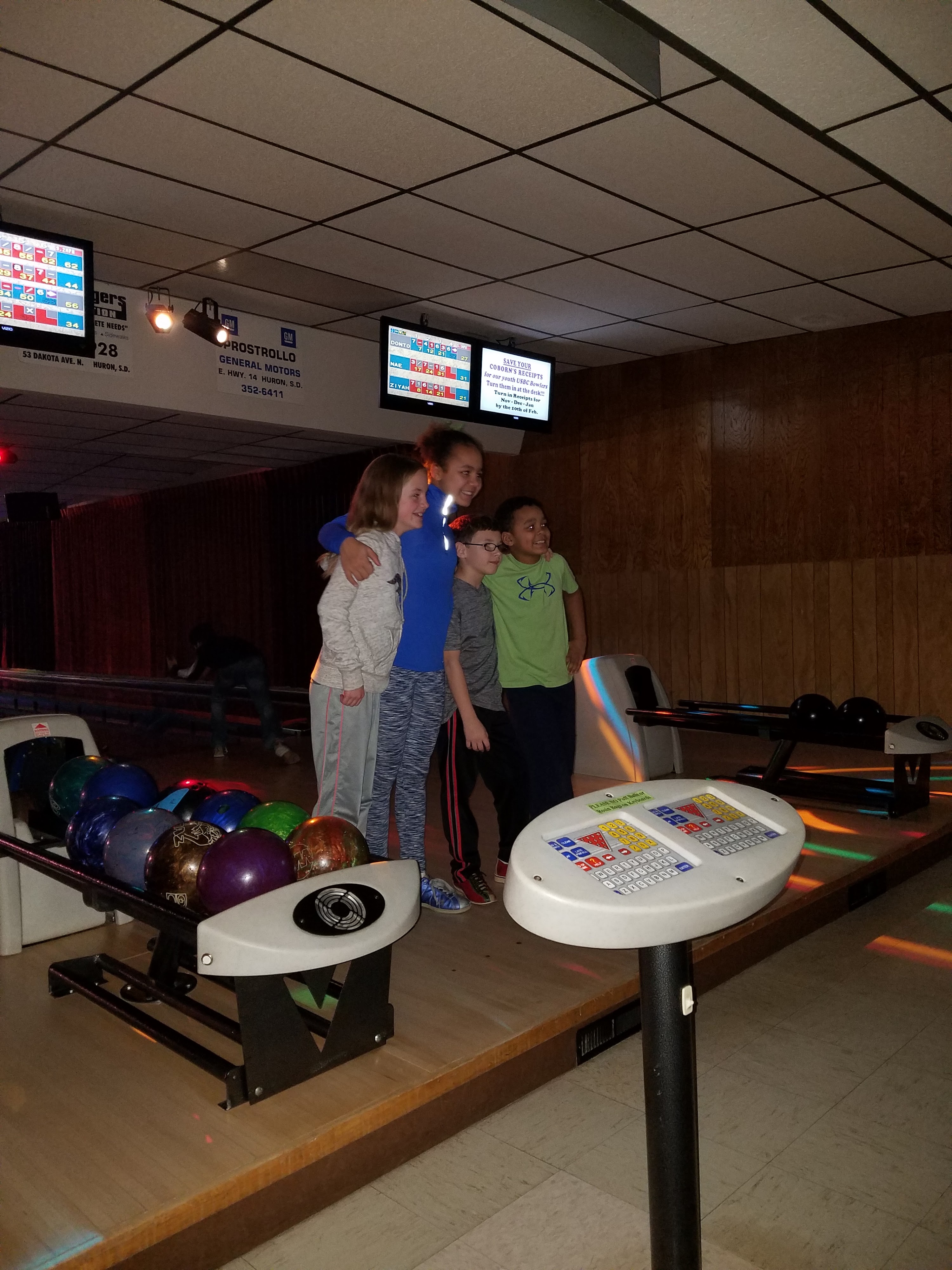 Four children pose at a bowling lane with colorful lights, a ball return, and automatic scoring consoles visible.