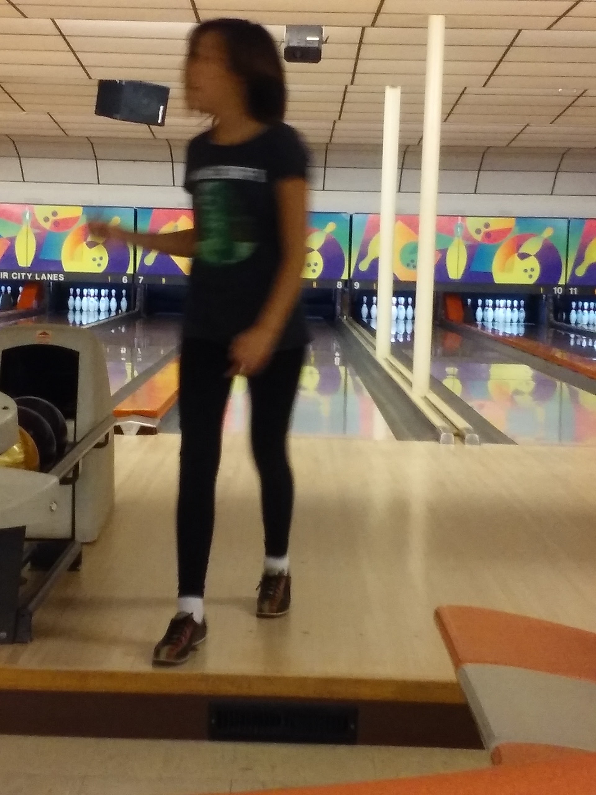 A young person in bowling shoes walks on an approach lane in a bowling alley, with multiple lanes and pins visible in the background.