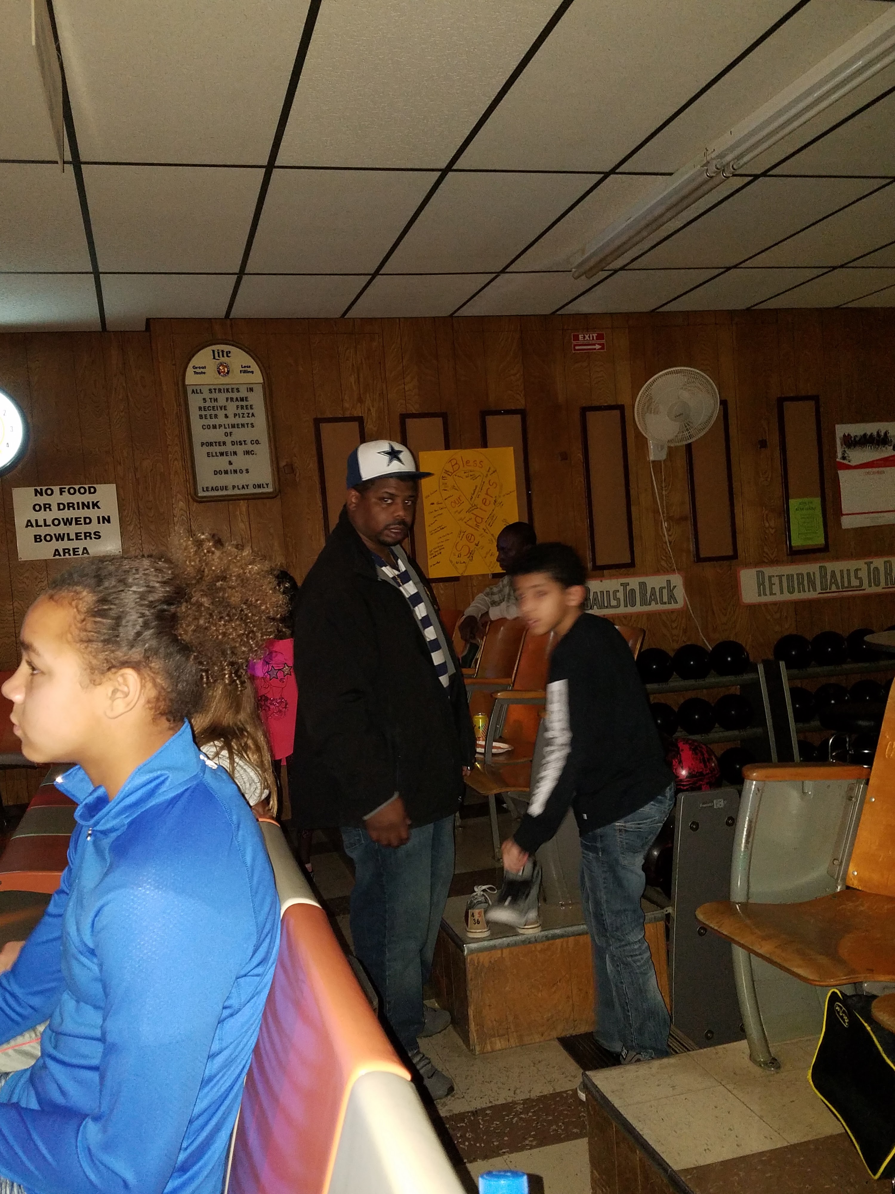 An adult and two children in a bowling alley, with bowling balls on a rack and wooden paneling in the background.