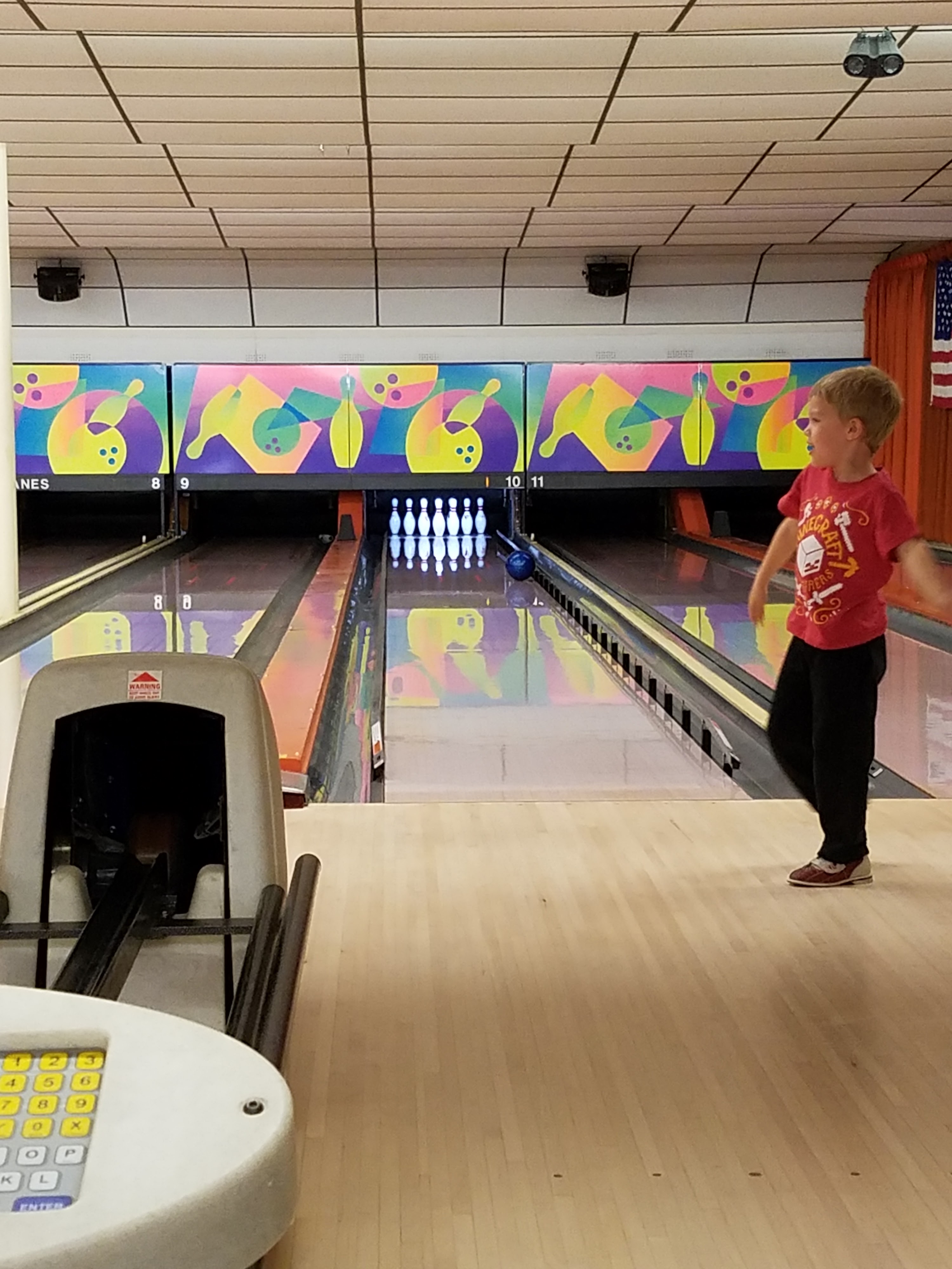 A young boy in a red shirt bowls a blue ball down a lane at a bowling alley.