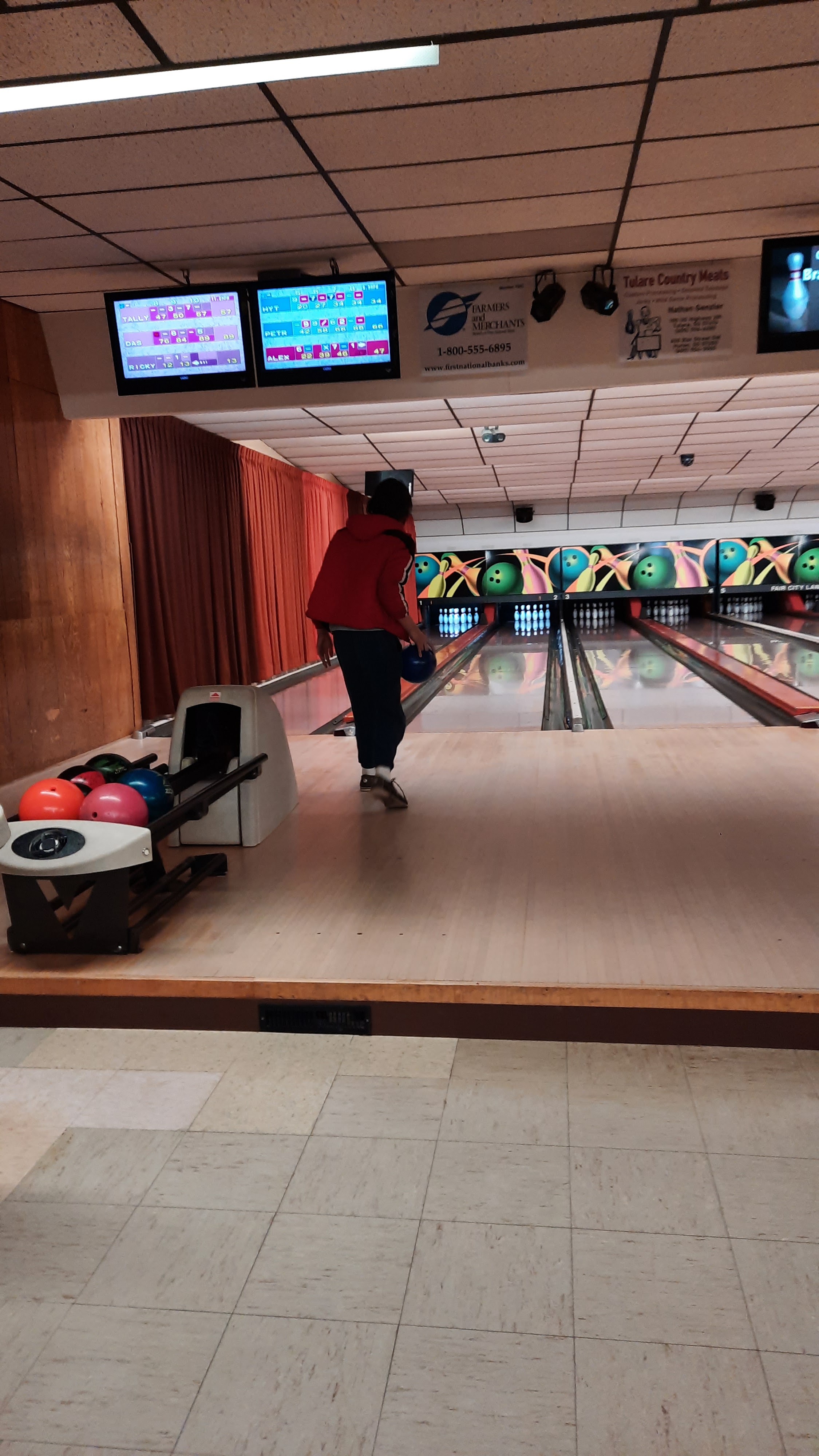 A person in a red hoodie bowls a blue bowling ball down a lane, with score screens overhead and multiple bowling balls in a return rack.