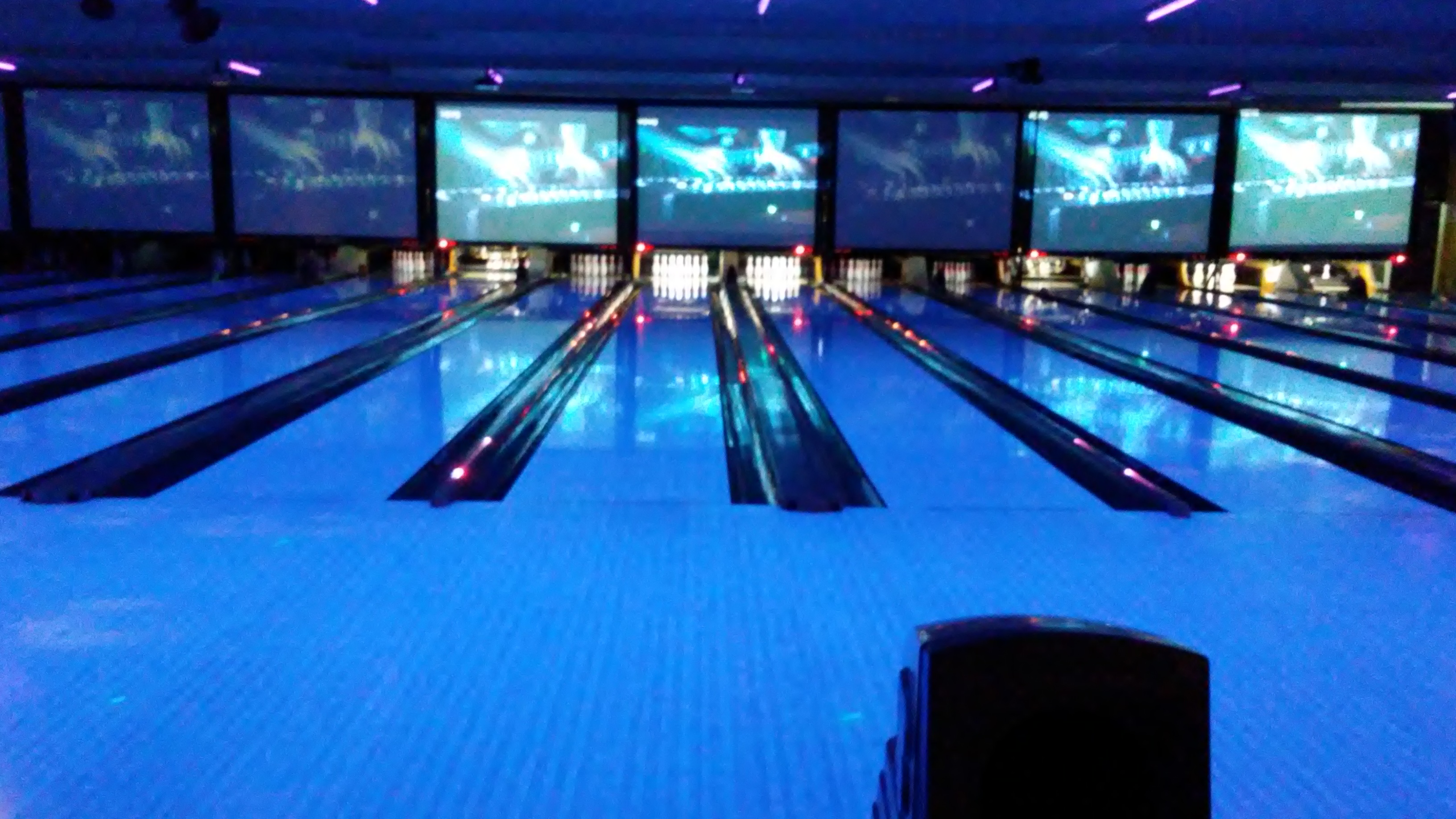 A view down several bowling lanes under glowing blue ultraviolet lights with large projection screens above the pins.