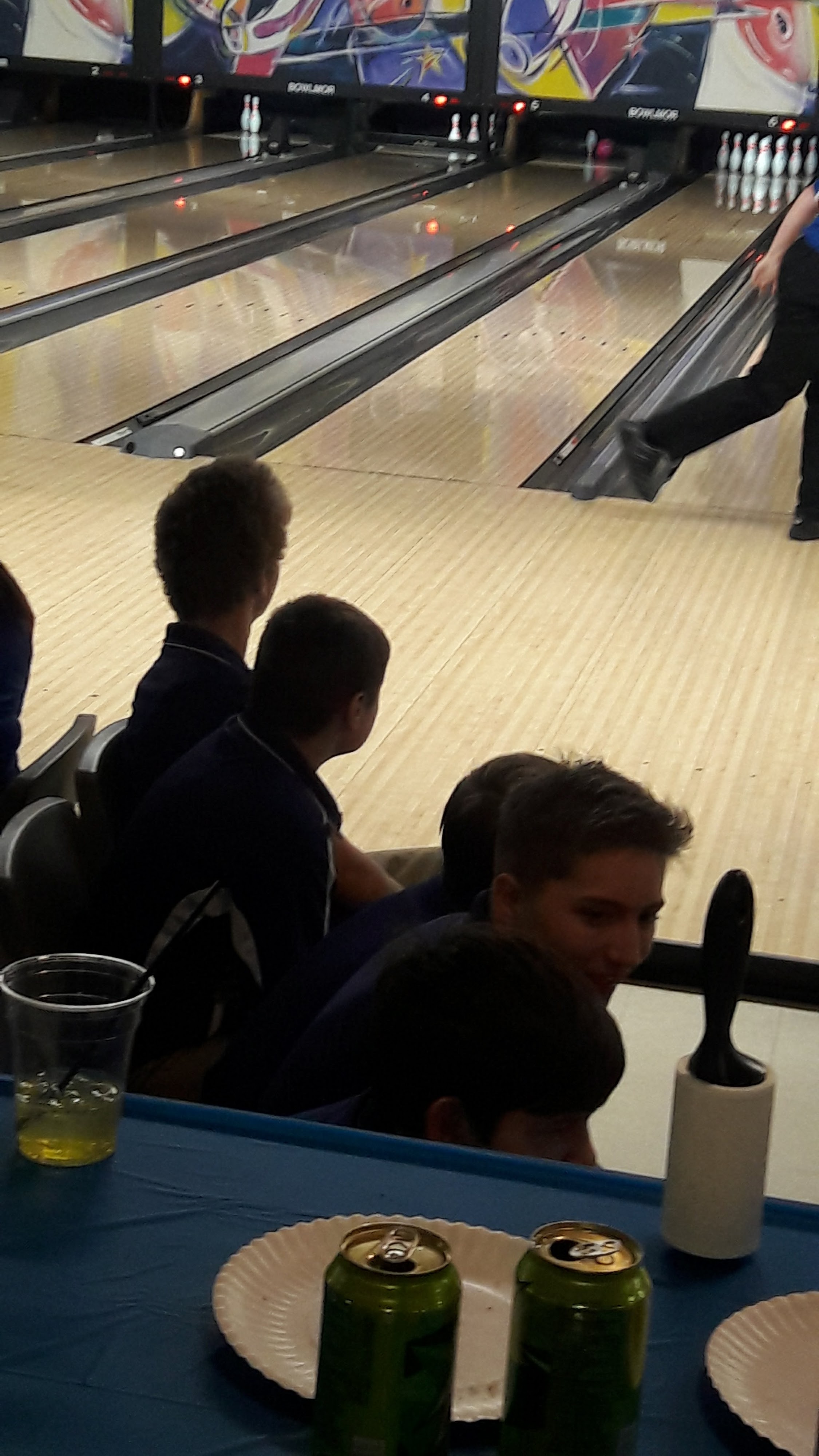 A group of young people sits near the bowling lanes with drinks on a table in the foreground.