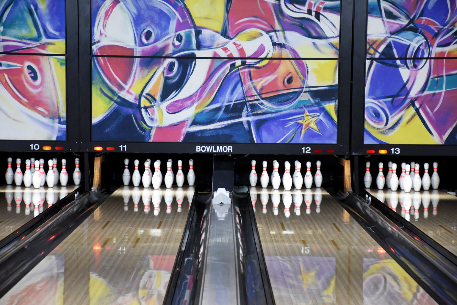 Four polished bowling lanes with full pin racks and colorful abstract masking units at Bowlmor Lanes.
