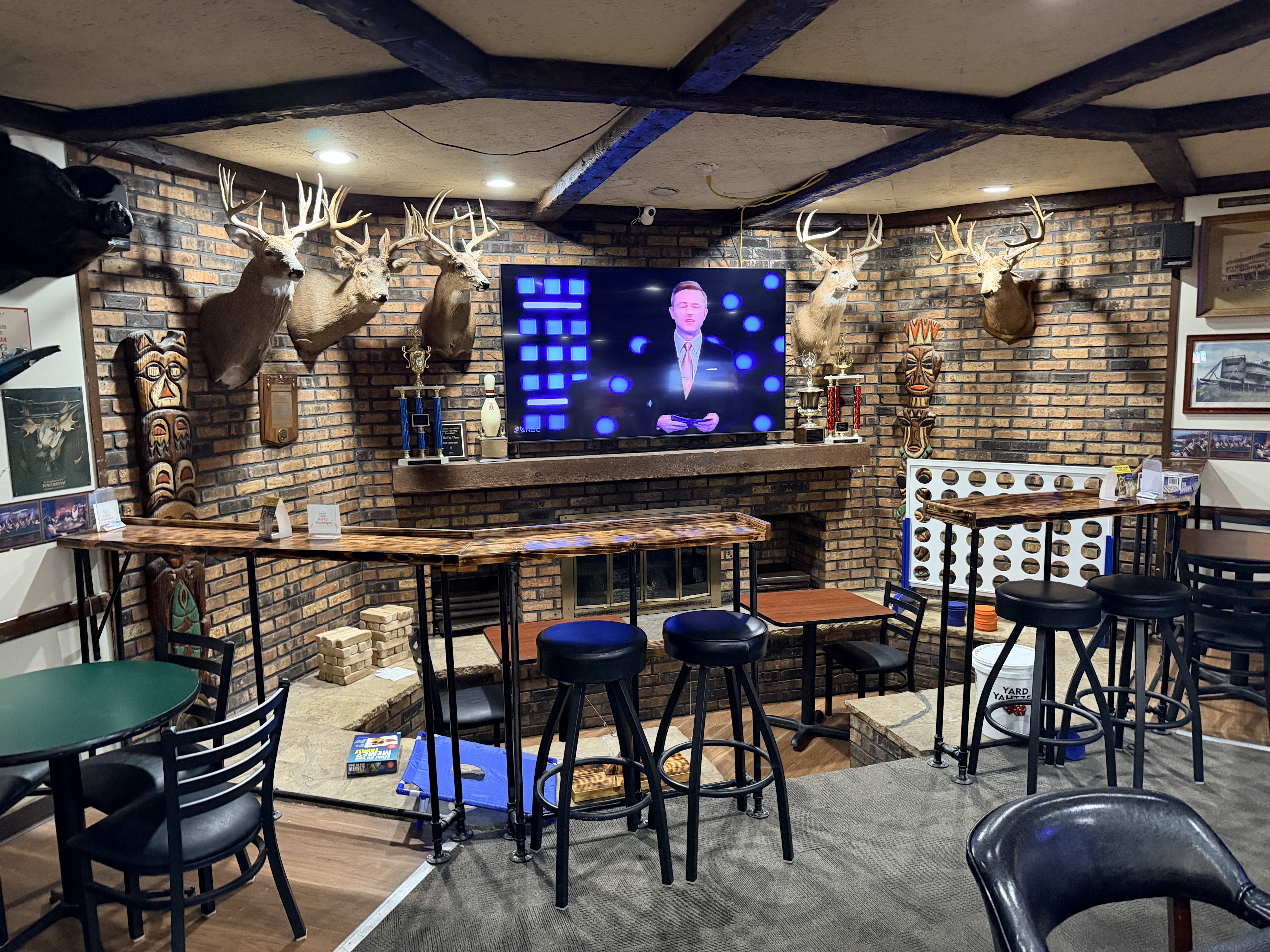 A rustic lounge with brick walls, taxidermy deer heads, high-top tables, and a large television.