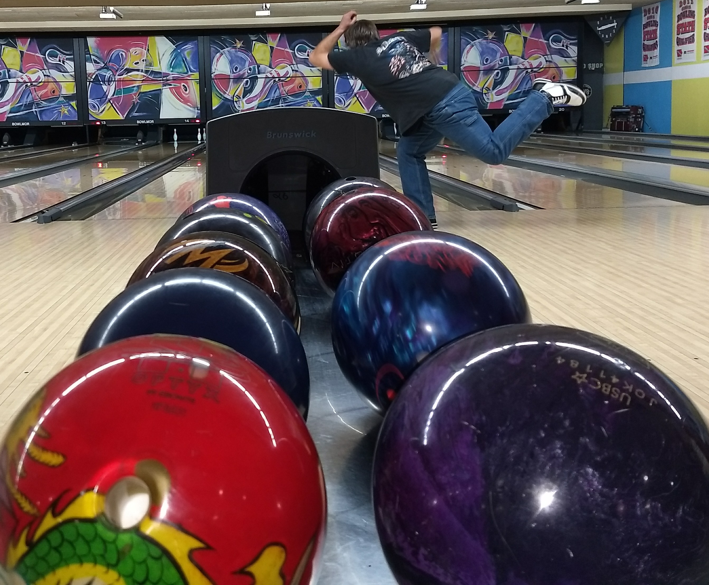 A low-angle view of colorful bowling balls on a return rack with a bowler in motion on a lane behind them.