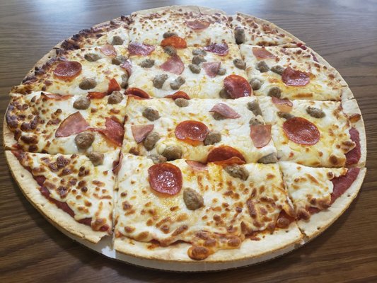 A thin-crust pizza with pepperoni, sausage, and ham cut into squares on a wooden table.