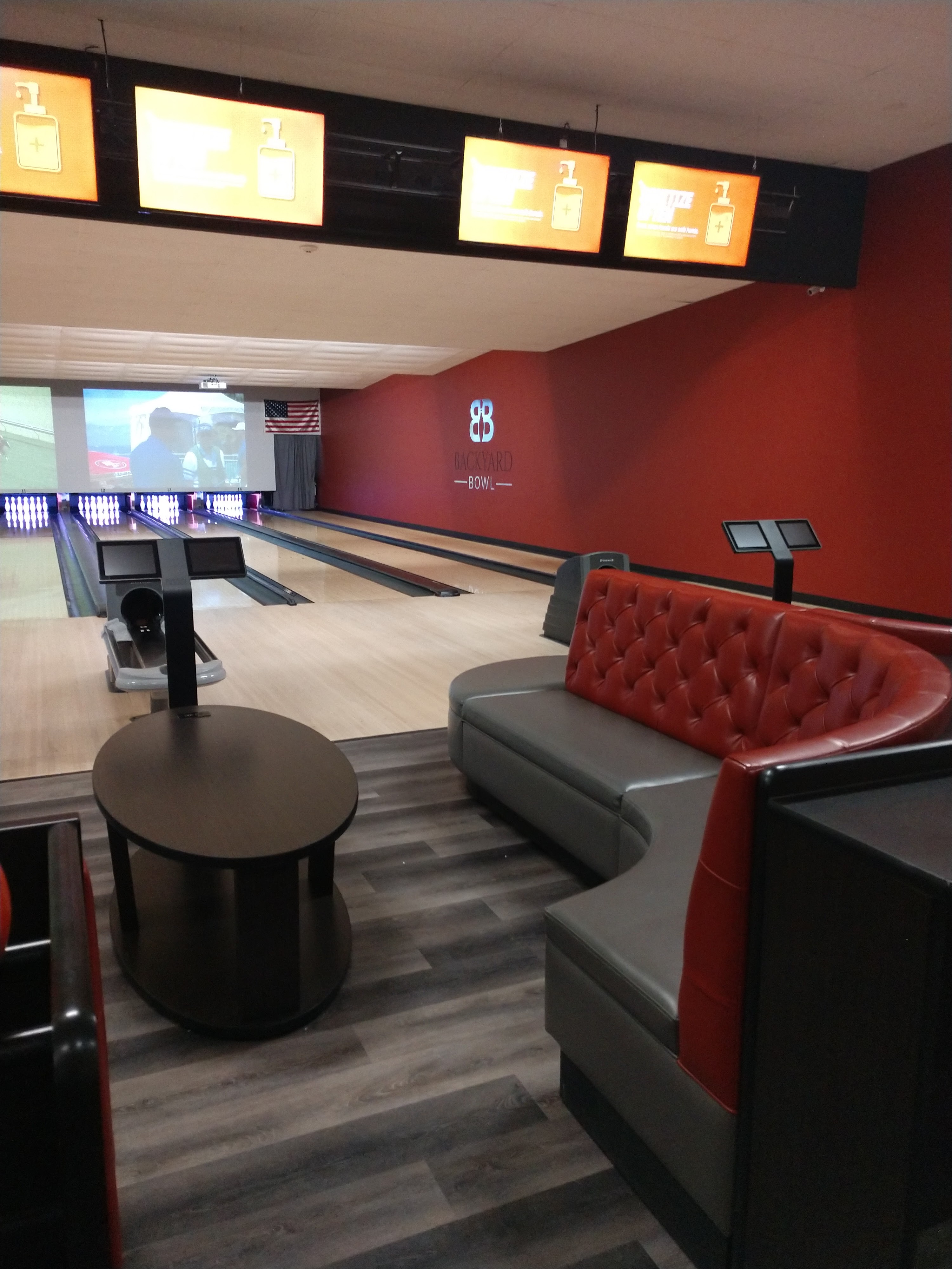 Interior of a modern bowling center with a red tufted couch, wooden lanes, and the Backyard Bowl logo on a red wall.