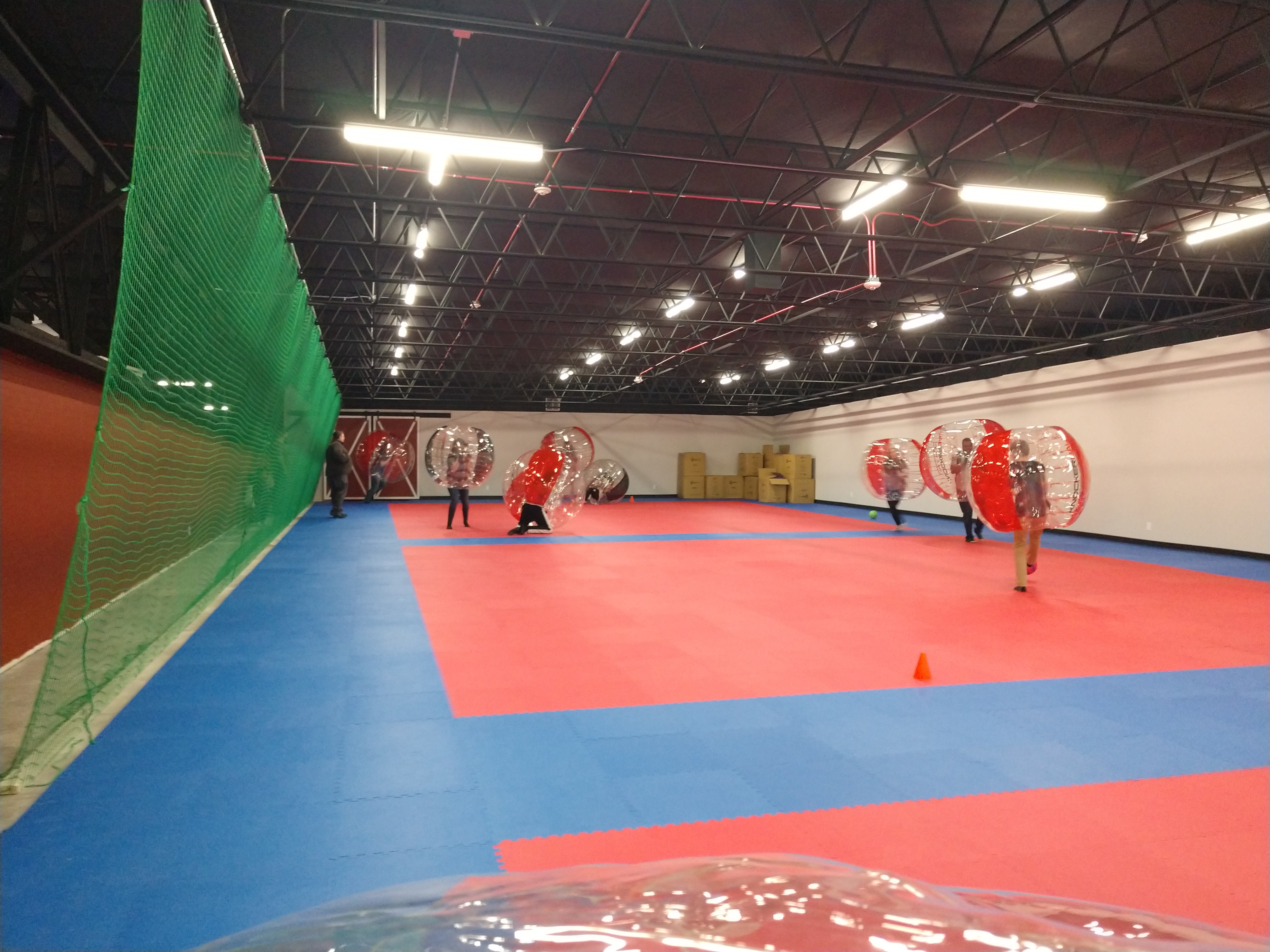 People in large inflatable bubble suits playing a game on a red and blue foam matted floor in an indoor arena.