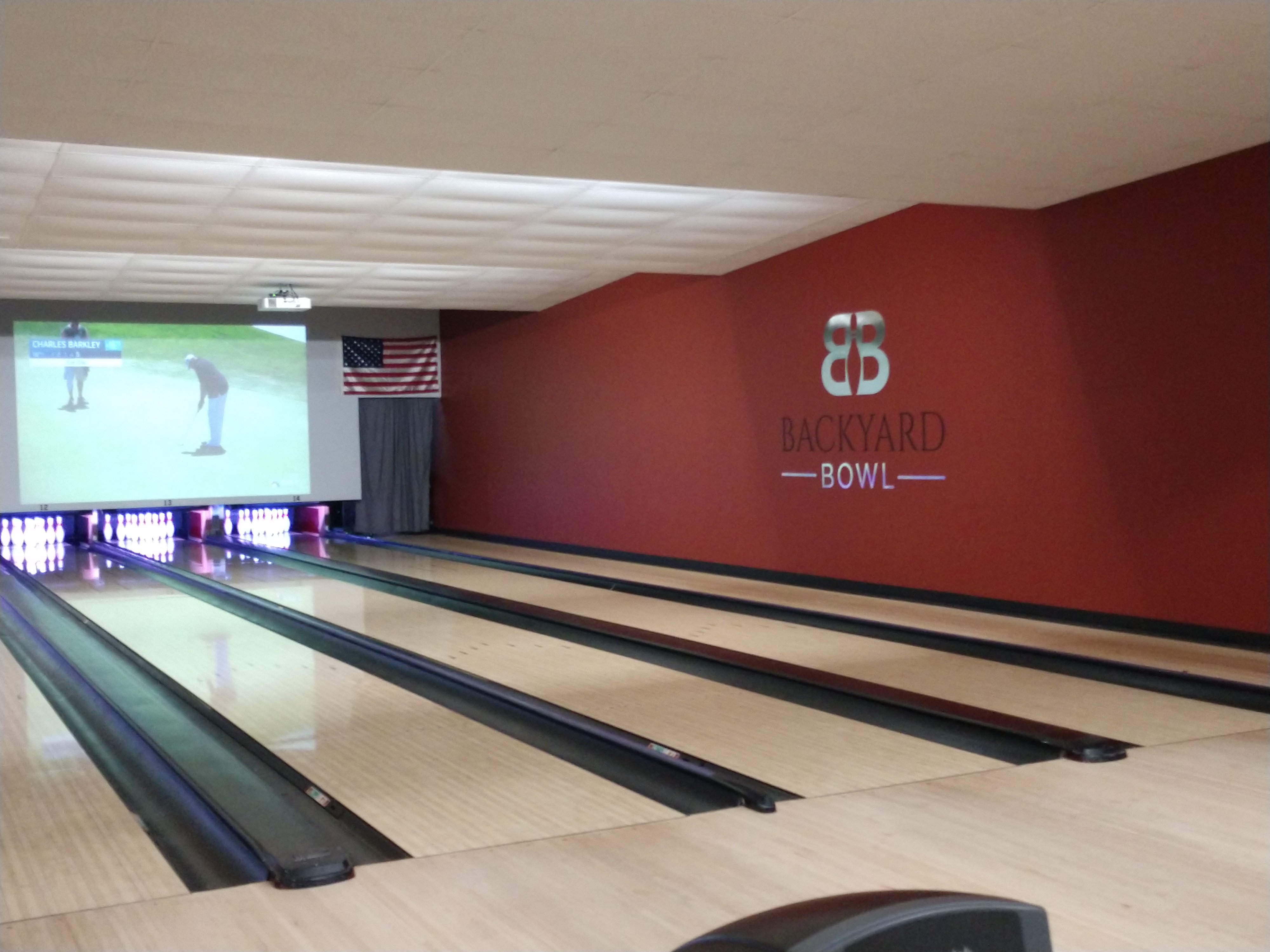 A view of modern bowling lanes next to a red wall featuring the Backyard Bowl logo and a large projection screen.
