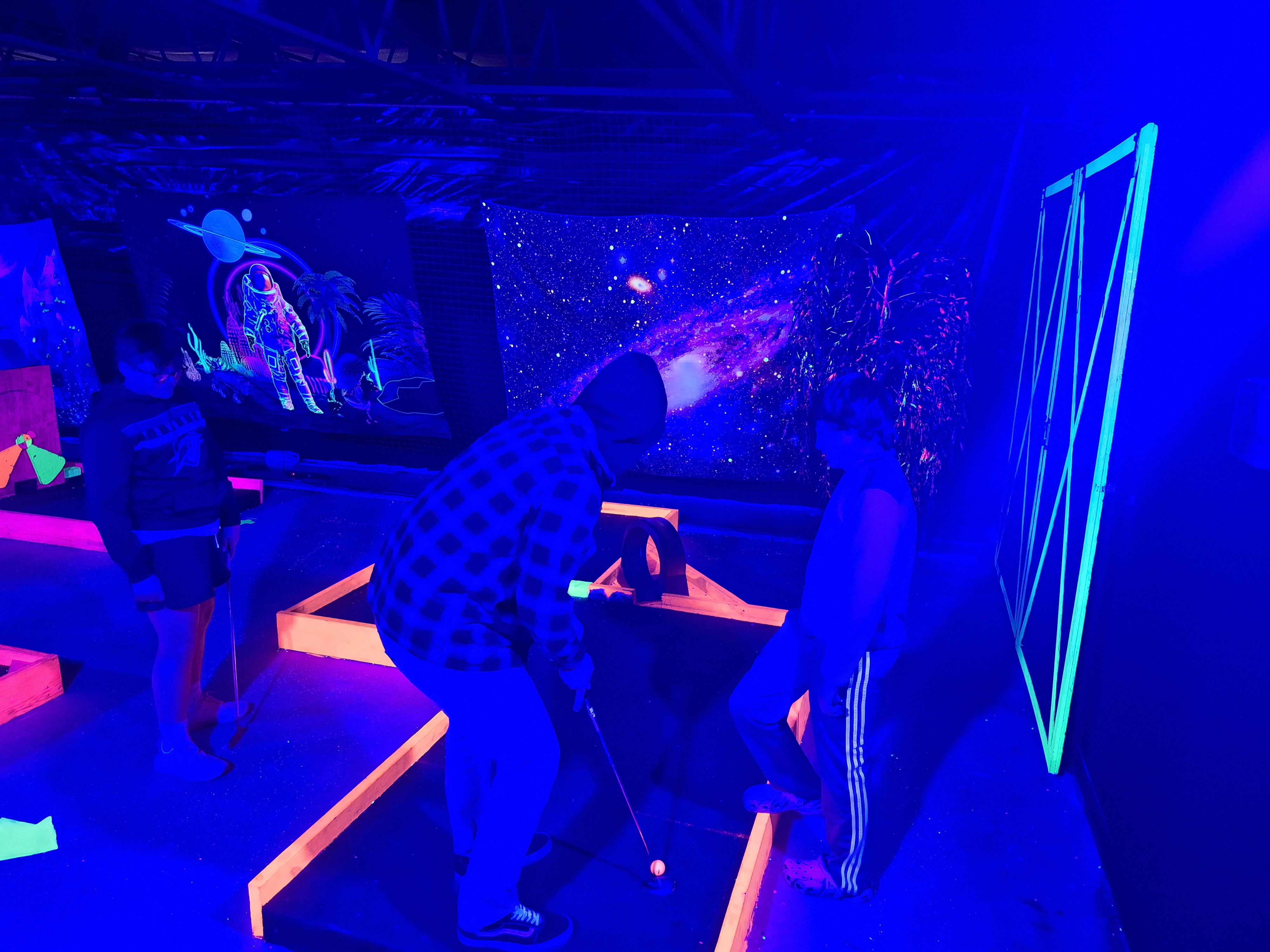People playing neon glow-in-the-dark mini-golf under purple UV lights with astronaut and space themed wall art.
