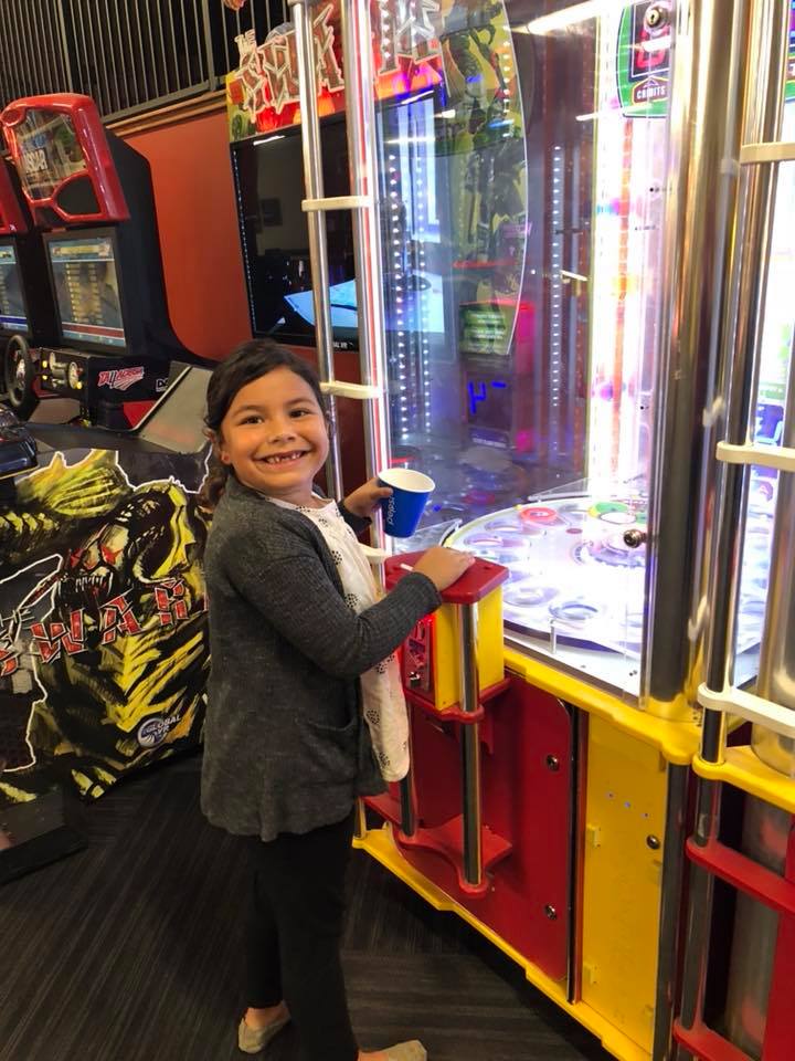 A young girl smiles while playing a tall, brightly lit arcade game in a game room.