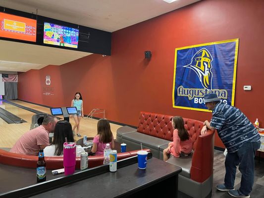 Families and friends relax in modern red tufted lounge seating at a bowling center with automatic scoring tablets.
