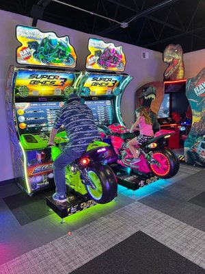 An adult and a child play a neon-lit motorcycle racing game in a modern arcade.