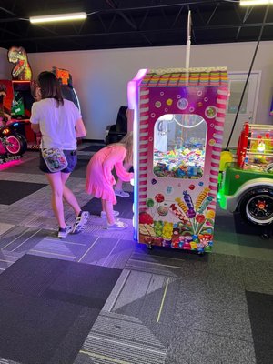 A child in a pink dress looks at a colorful candy claw machine in an arcade with an adult standing nearby.