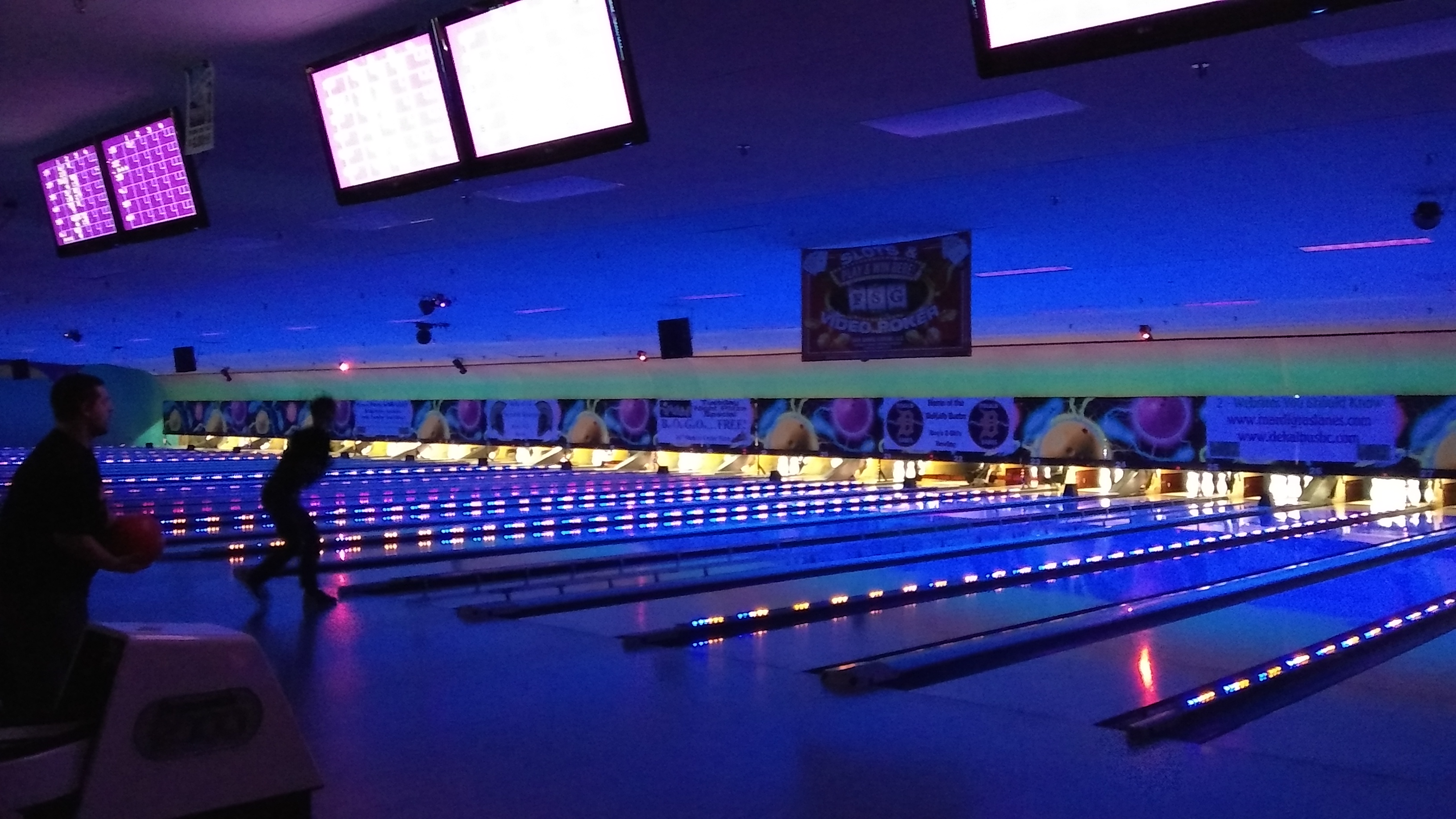 People bowling at Mardi Gras Lanes during a cosmic session with neon blue lights and glowing lane markers.