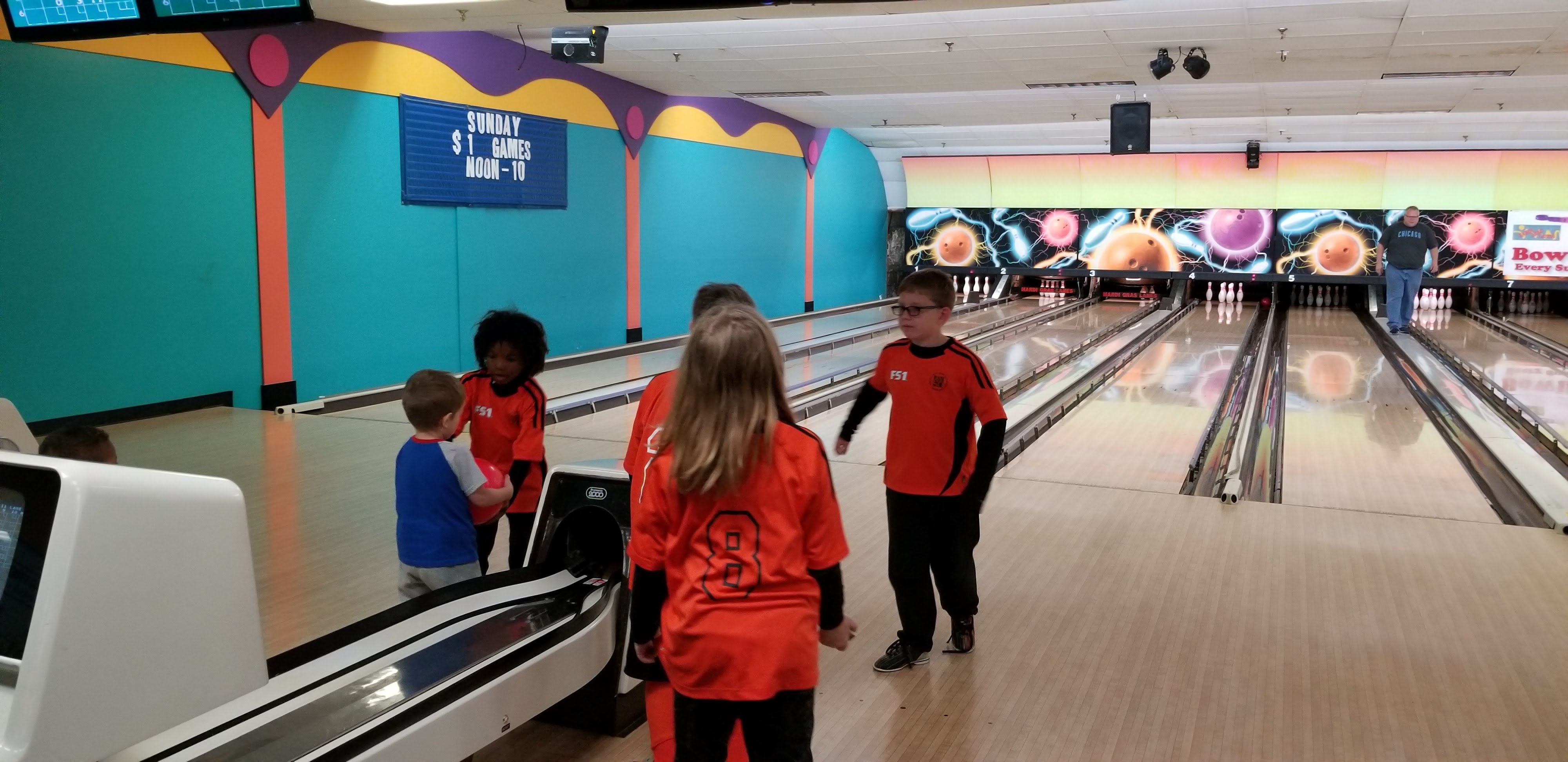 Children in orange jerseys gather at the bowling lanes next to a ball return.