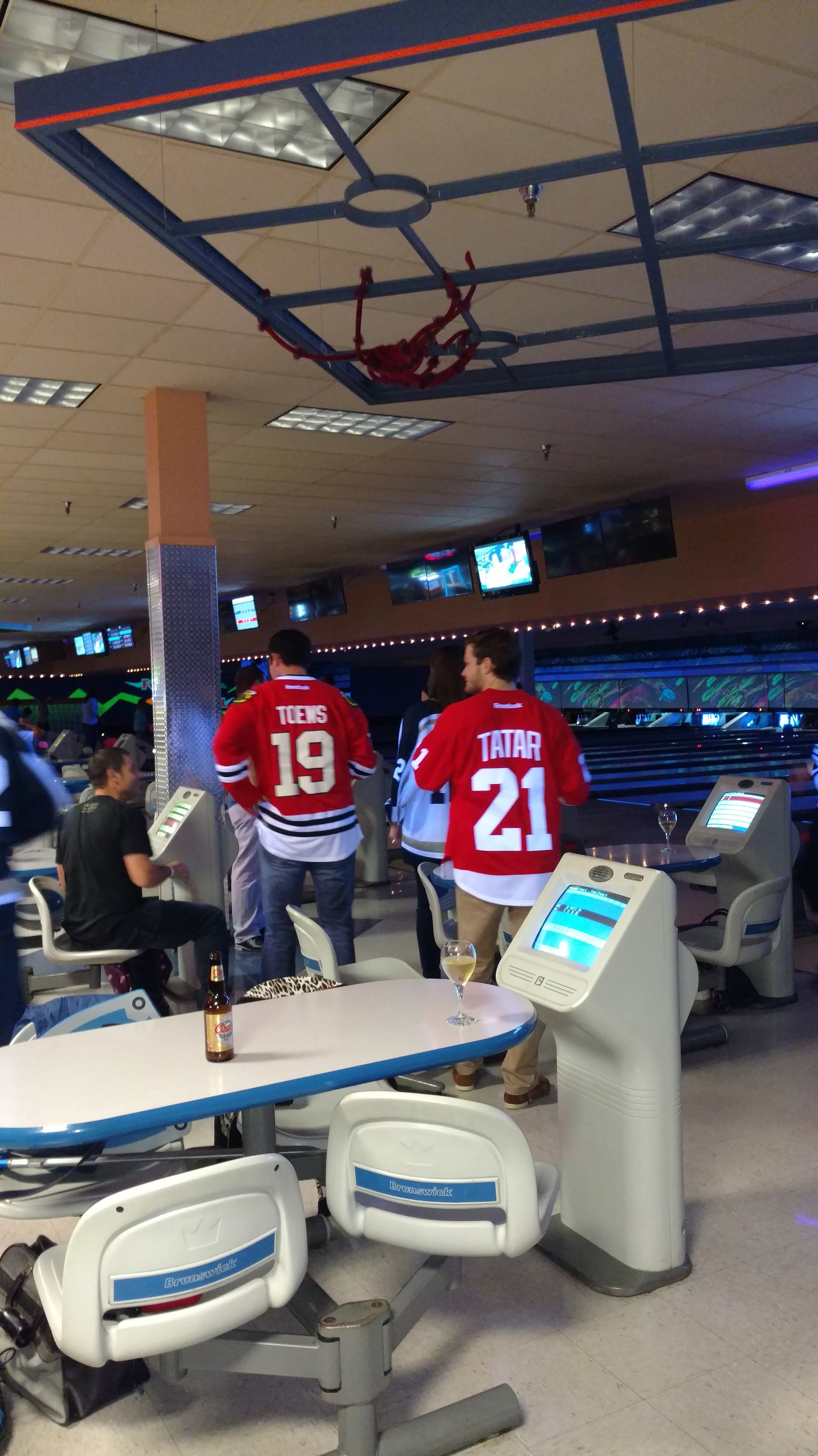 A group of people in hockey jerseys standing in a bowling alley settee area near lanes and scoring consoles.
