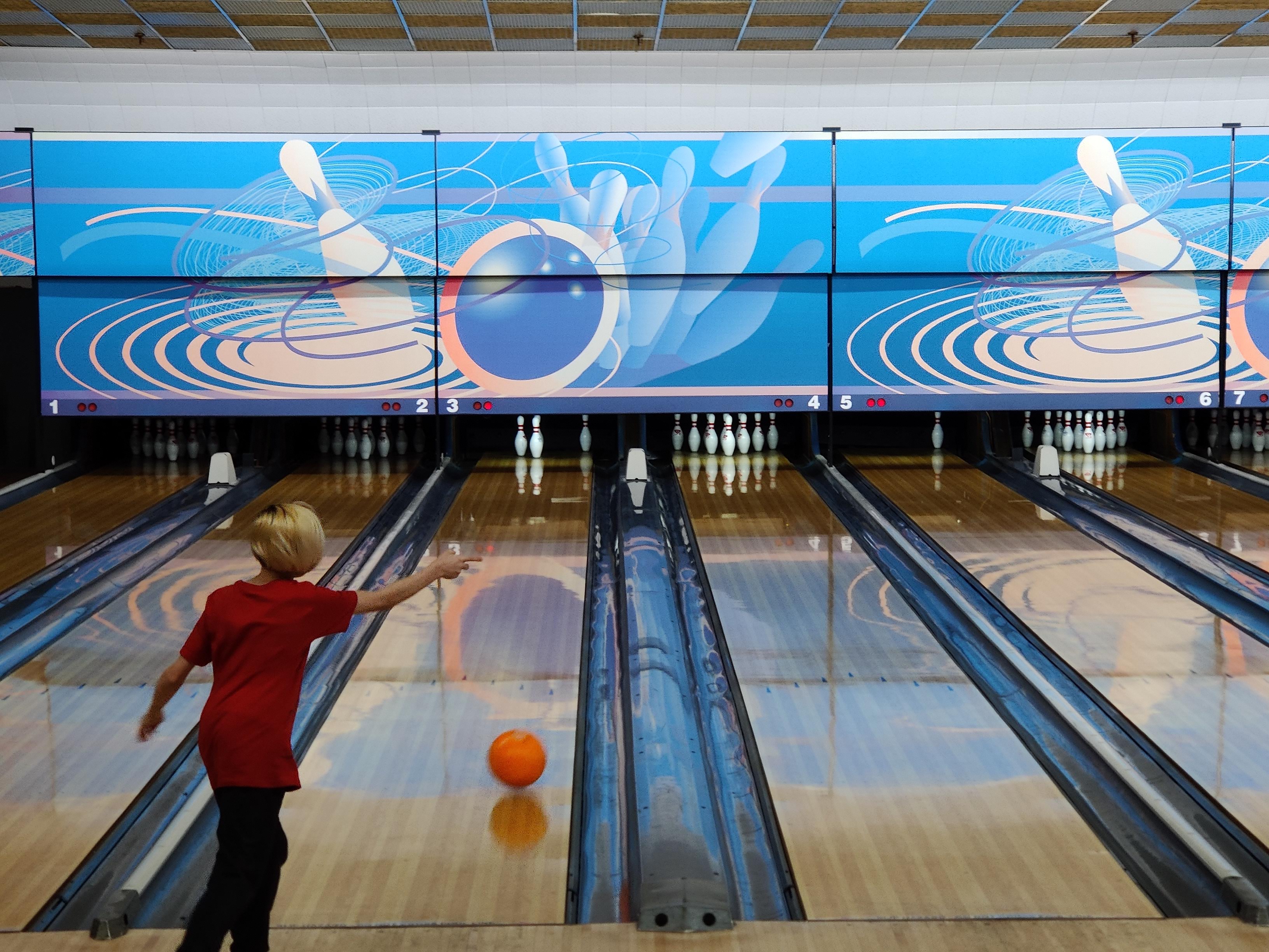 A young person in a red shirt bowls an orange ball down a lane at Fox Bowl.