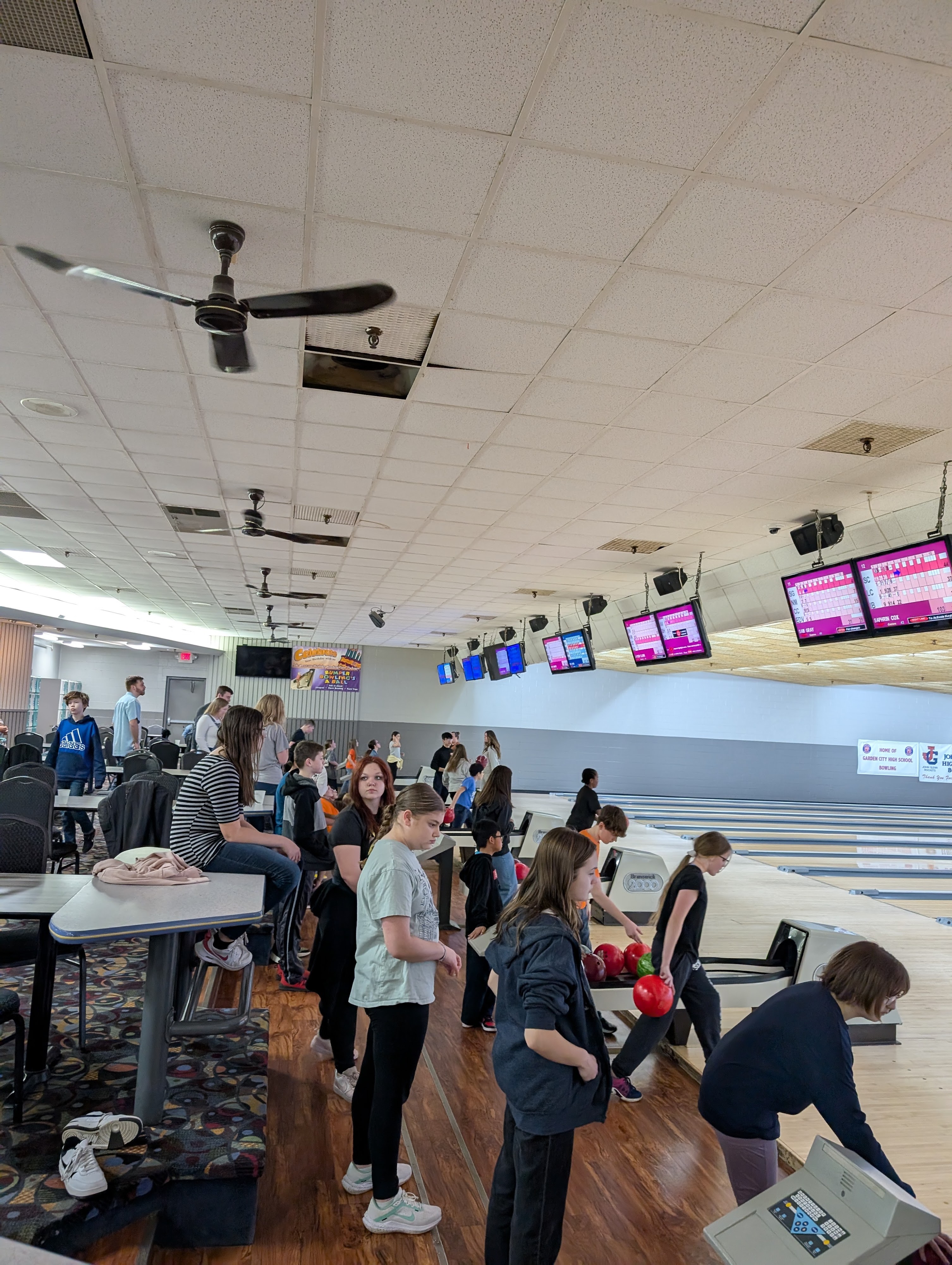 A large group of teenagers and children bowling in a well-lit center with wooden lanes and overhead scoreboards.