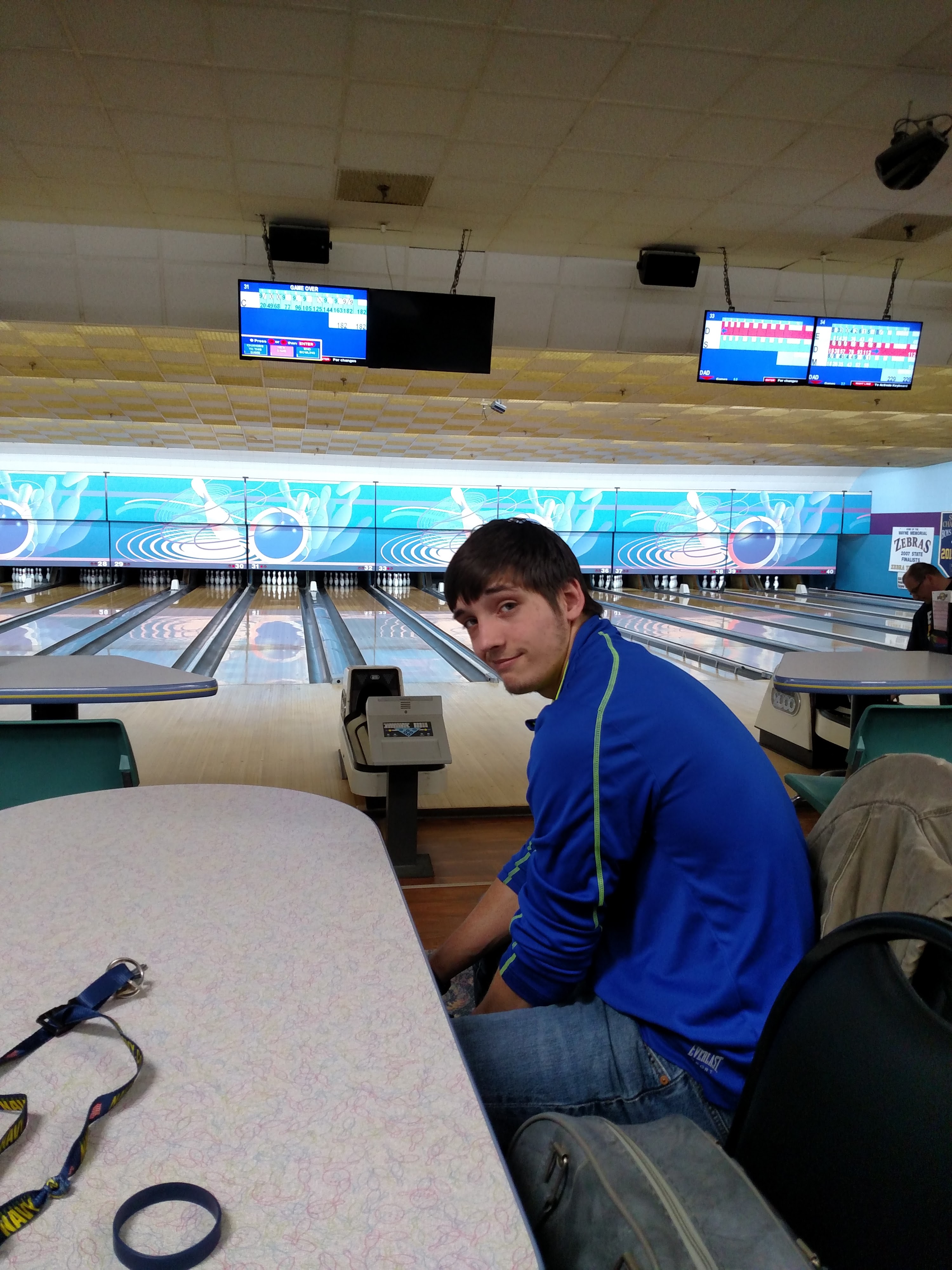 A young man in a blue jacket sits at a table in a bowling center with lanes and scoring monitors in the background.