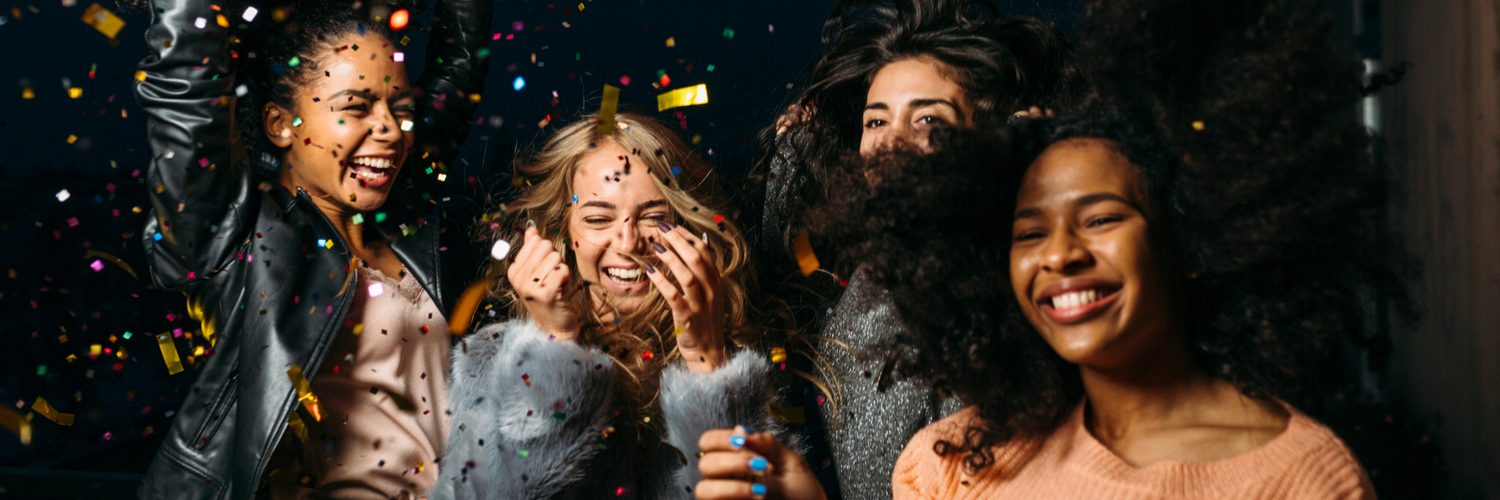 Four women laugh and celebrate under falling colorful confetti against a dark background.