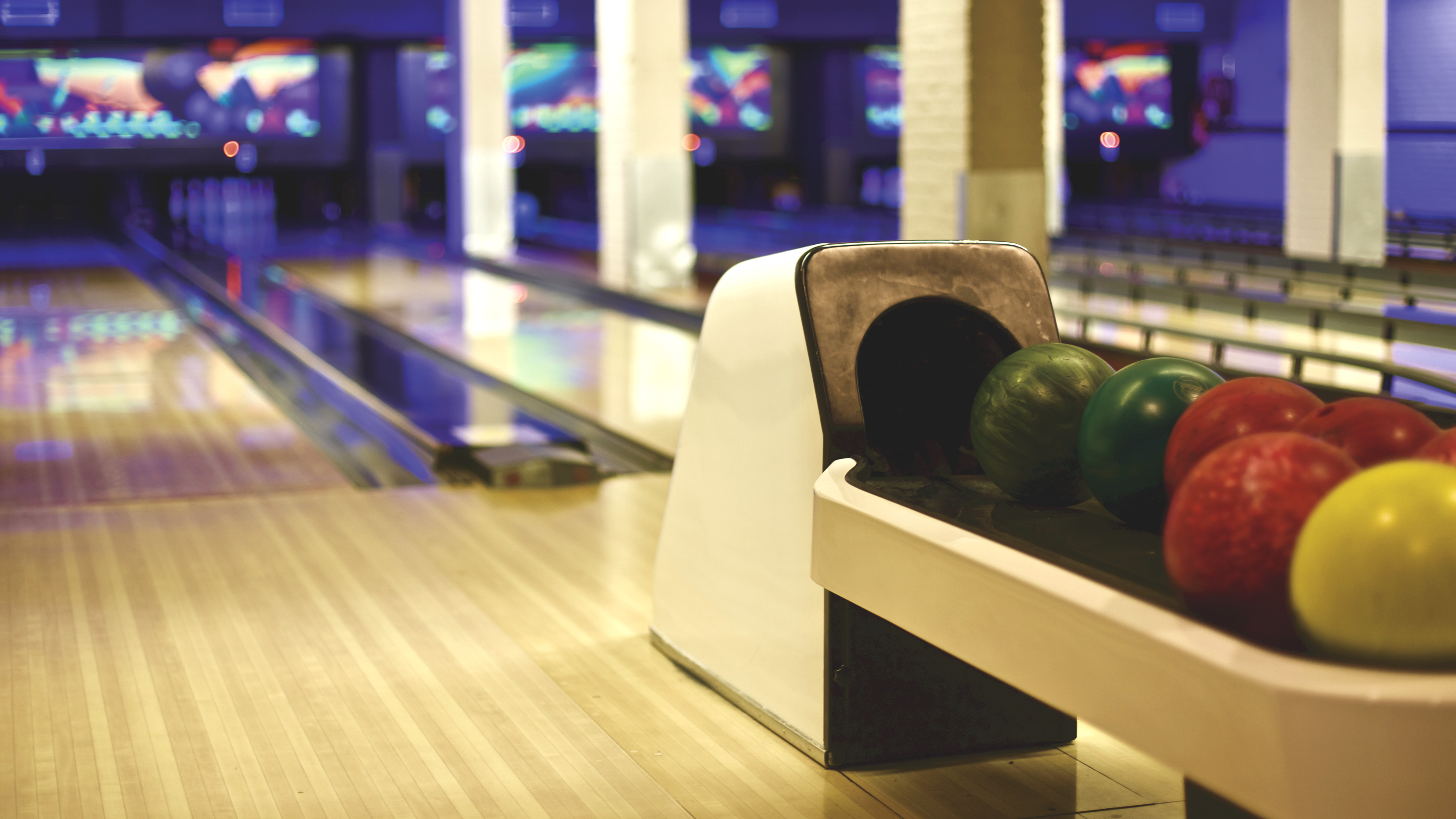 Close-up of green, red, and yellow bowling balls on a white return with blurred lanes in the background.