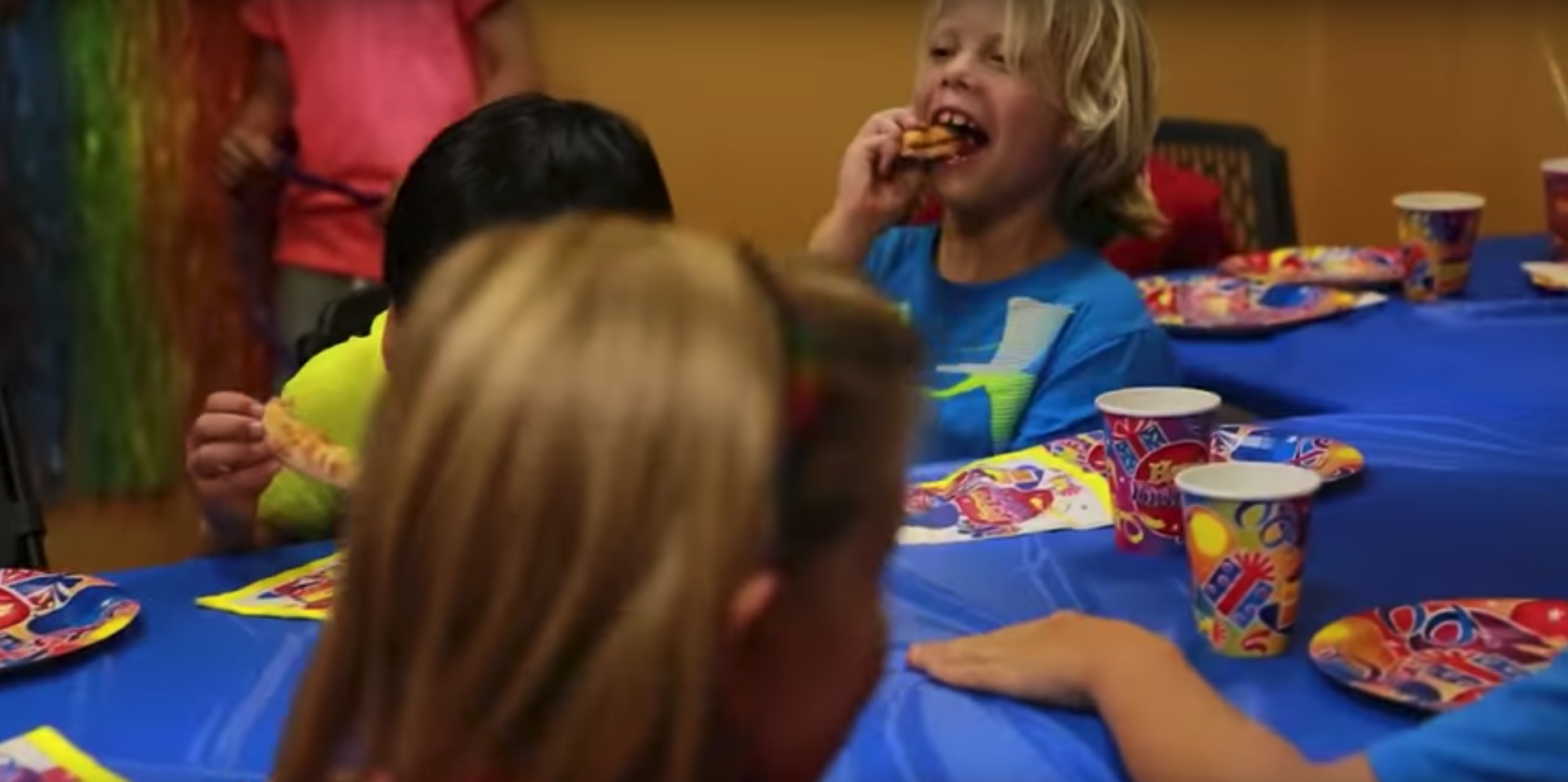 Children sitting at a blue table eating pizza and celebrating a birthday party.