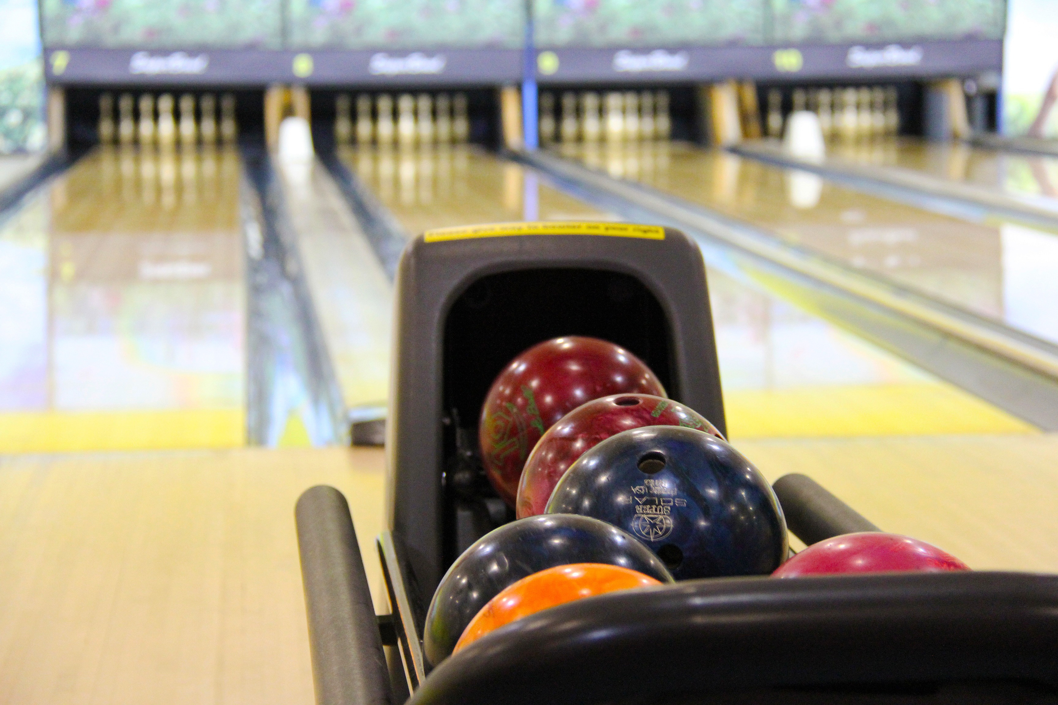 Close-up of colorful bowling balls in a ball return rack with blurred bowling lanes in the background.