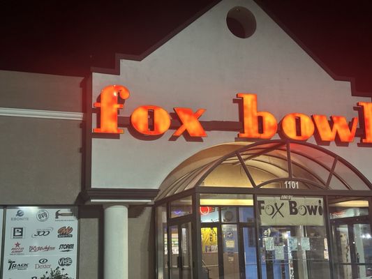 The exterior entrance of Fox Bowl at night with an illuminated orange sign and glass doors.