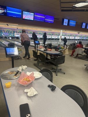 Patrons bowling at a center with snacks on a table in the foreground.