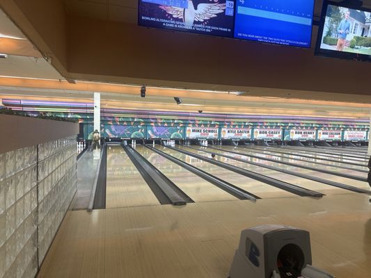 A view of multiple bowling lanes with overhead scoring monitors and names of 300-game bowlers above the pins.