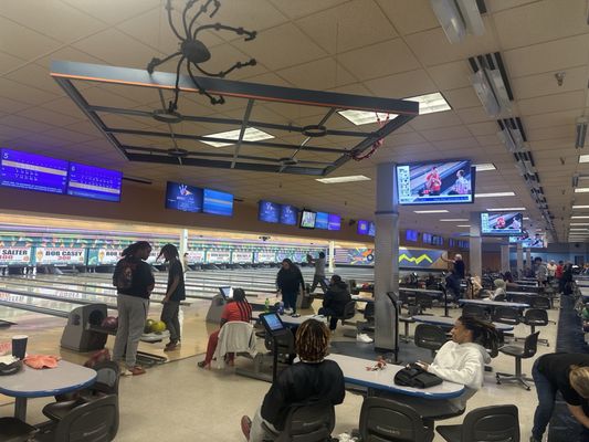 A busy bowling alley with multiple lanes, overhead monitors, and people enjoying a game from the seating area.