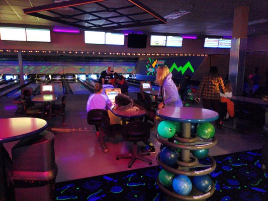 A bowling center with neon cosmic lighting, active lanes, and groups of people socializing in the settee area.