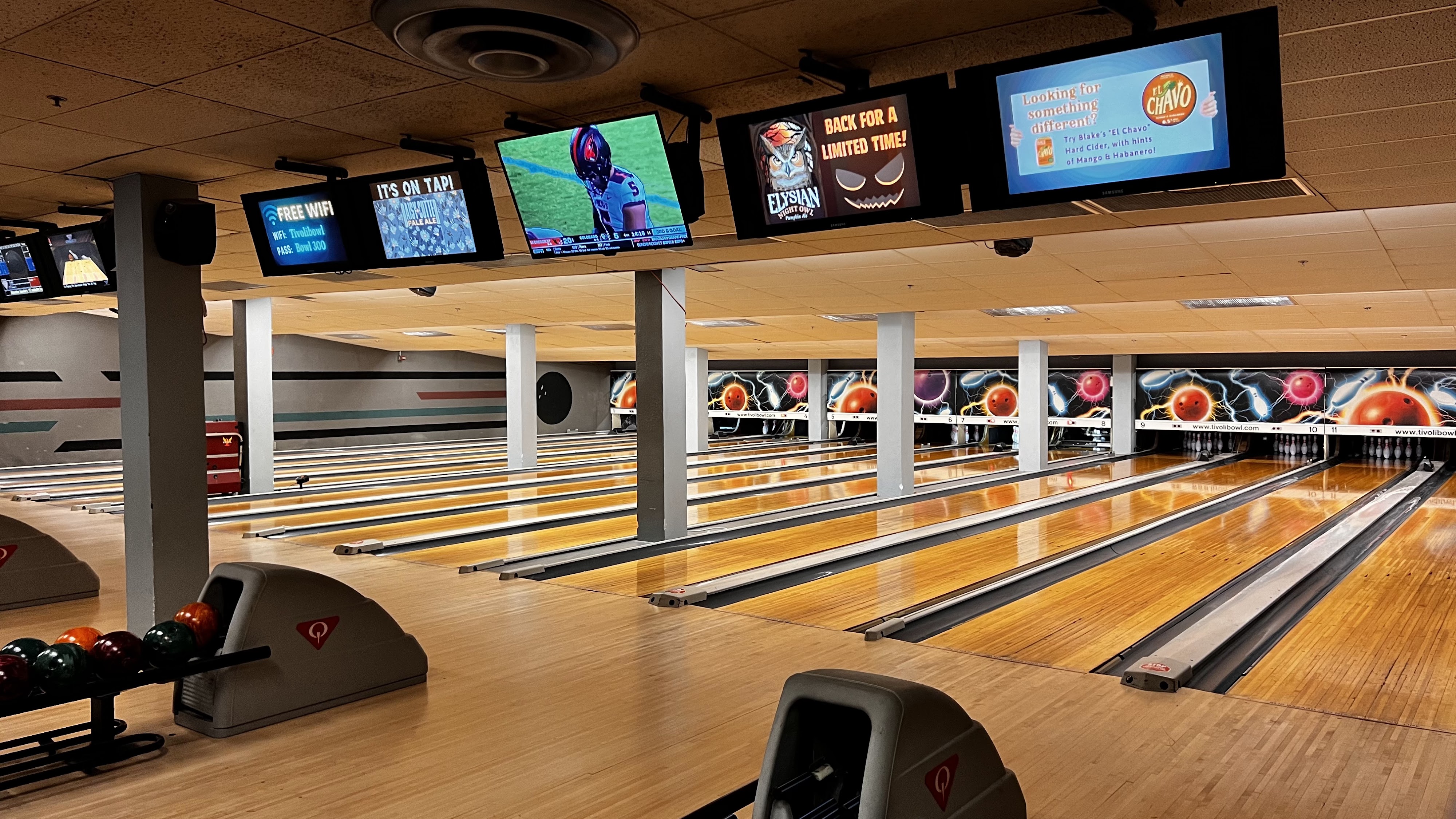 A wide view of polished wooden bowling lanes with overhead monitors and colorful back wall graphics.