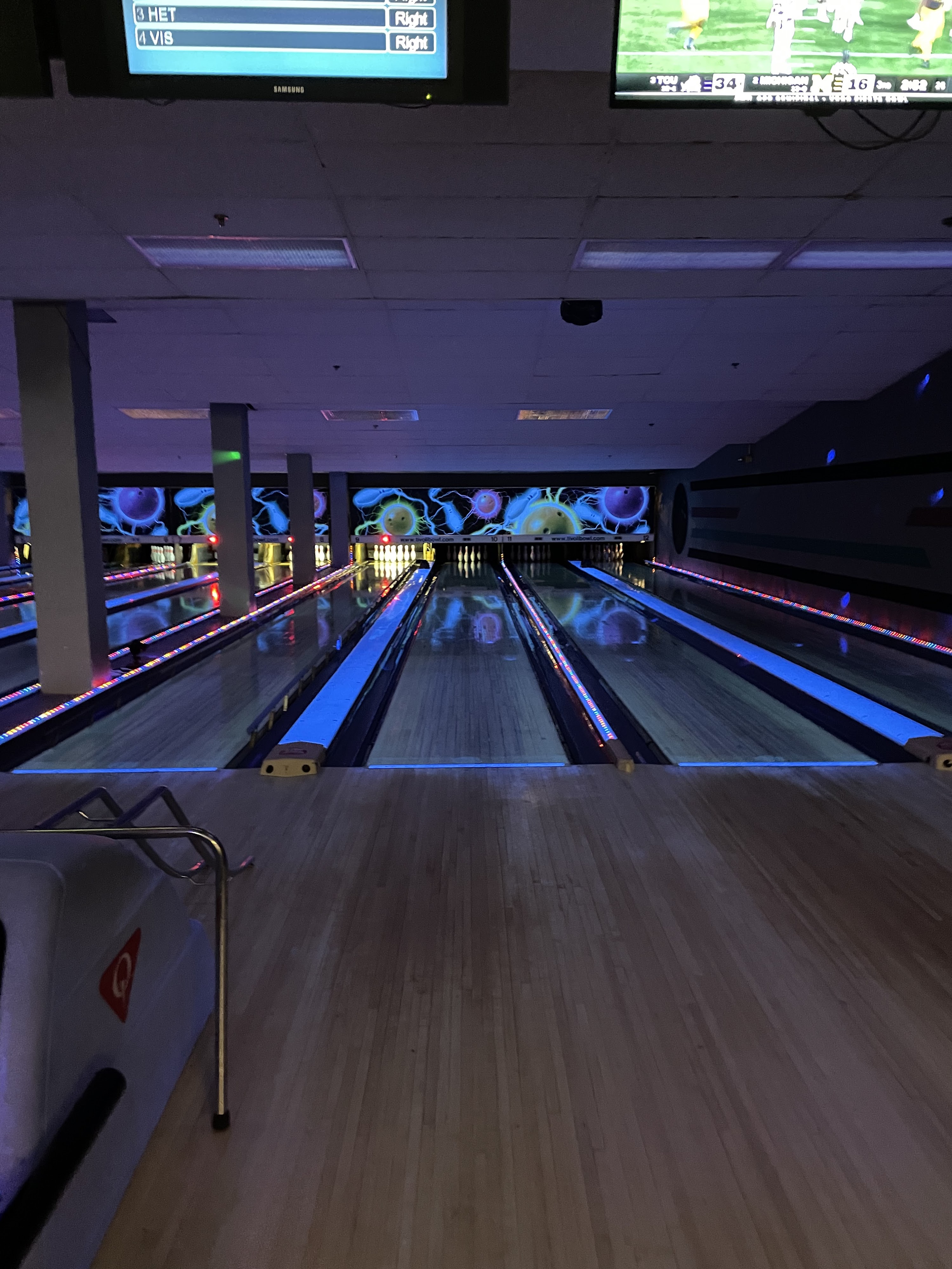 A vertical view of bowling lanes with bright blue LED gutter lighting and cosmic murals under overhead monitors.